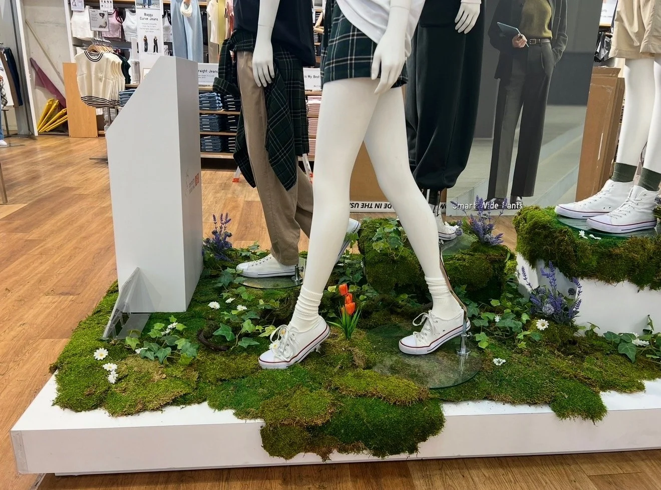 Display of mannequins in a clothing store, featuring one mannequin with white legs dressed in white sneakers and socks, standing on simulated grass and moss with small flowers, surrounded by other mannequins in pants and shirts.