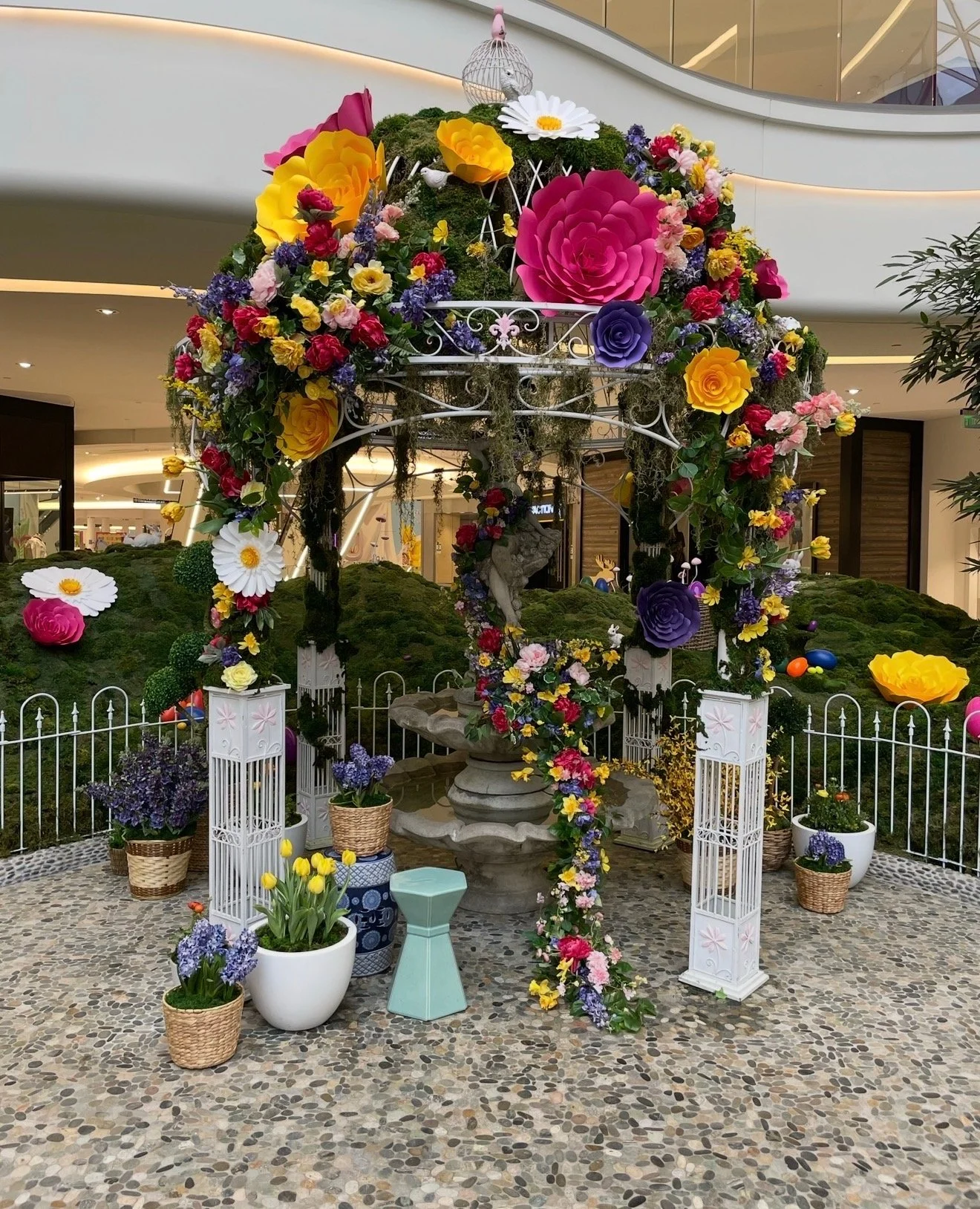 A decorative floral display with various colorful paper flowers, greenery, and small sculptures set up in an indoor shopping mall. The display is surrounded by potted plants and is enclosed by a small white fence.