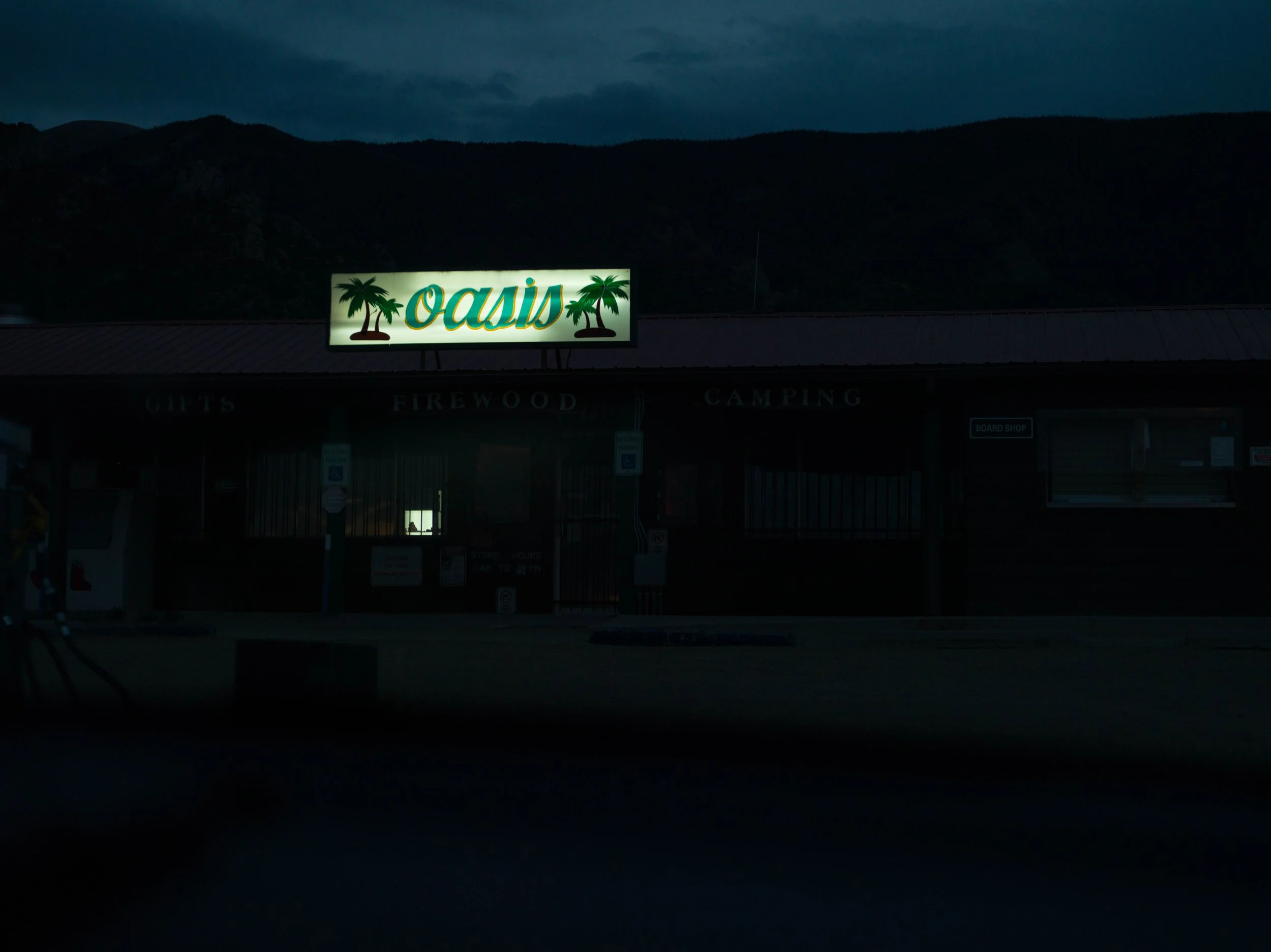 A lit sign on a building reads 'OASIS' with palm tree graphics, with a mountainous background at dusk or night.