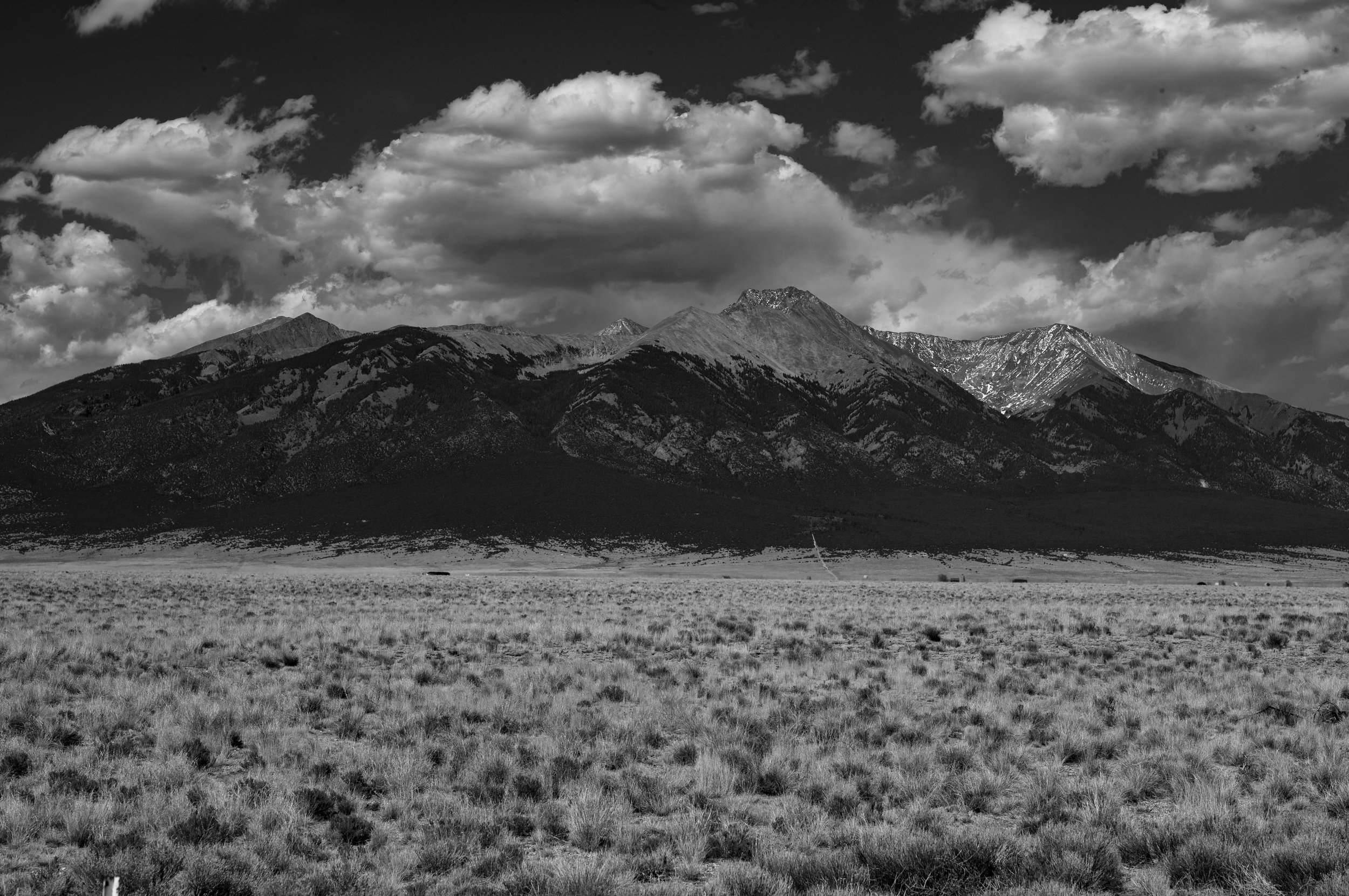 Black and white image of a mountain range with clouds overhead and an open grassy plain in the foreground.