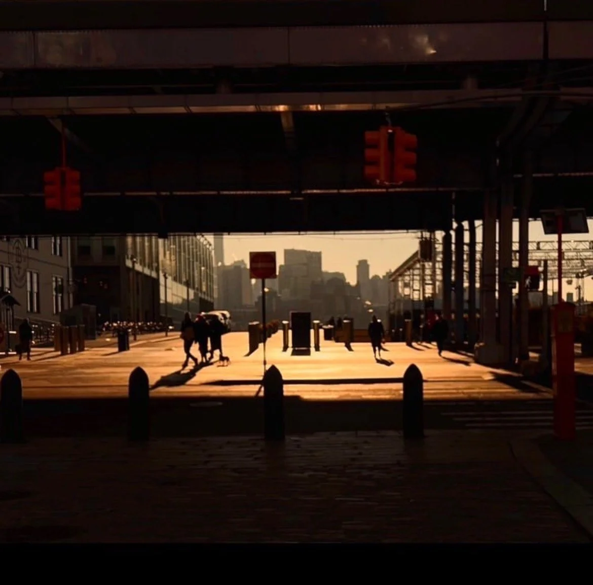 Street scene during sunset with silhouettes of people walking, buildings in background, and traffic lights overhead.