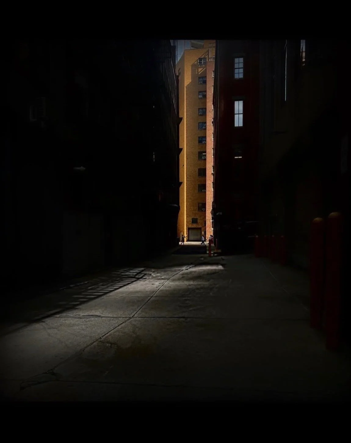 A dark alley leading to a brightly lit street with tall buildings and people in the background.