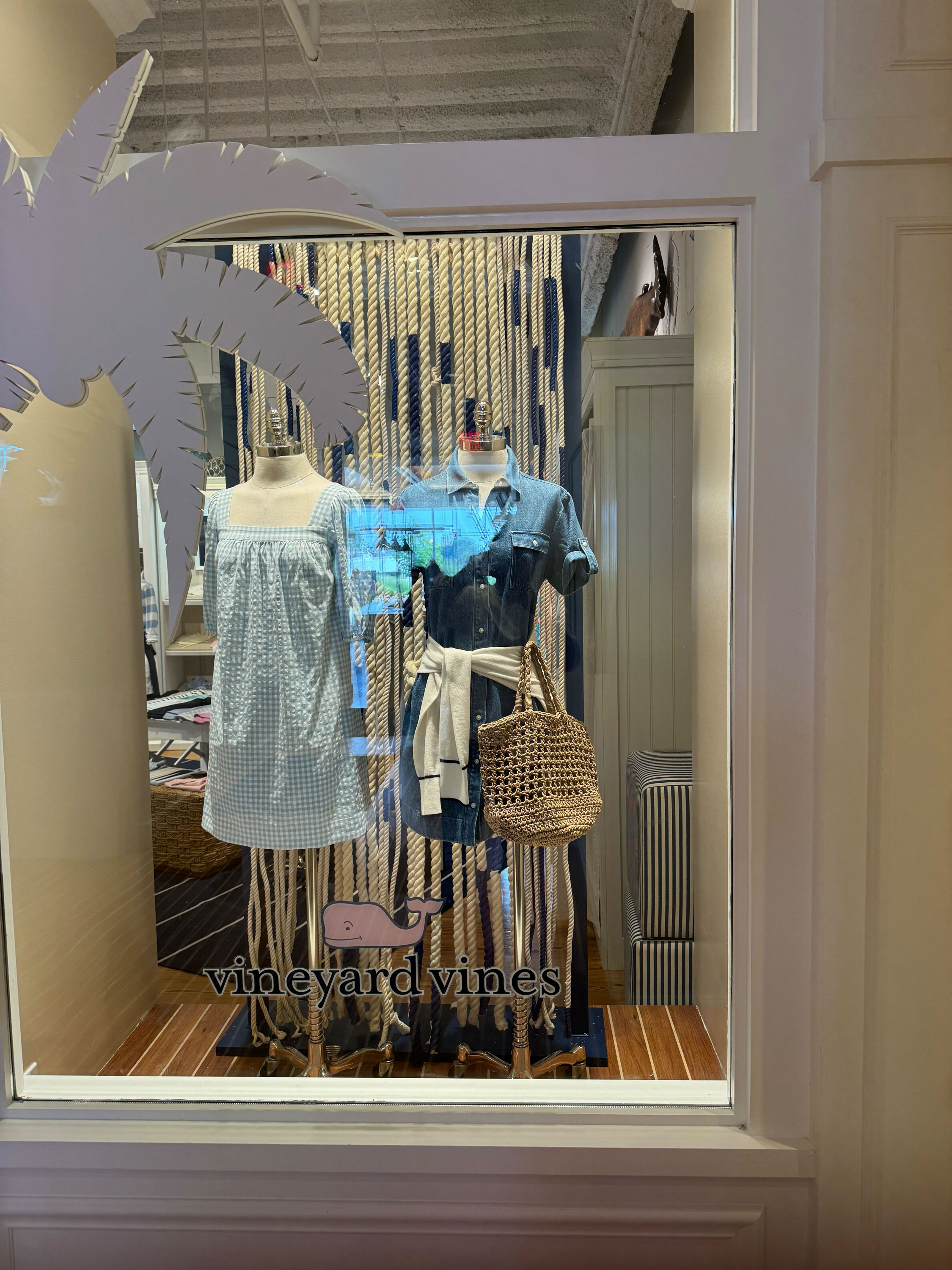 Store window display with mannequins wearing casual clothing, a sign with a pig illustration and the text 'vineyard vines' at the bottom, and a background with vertical rope decoration.