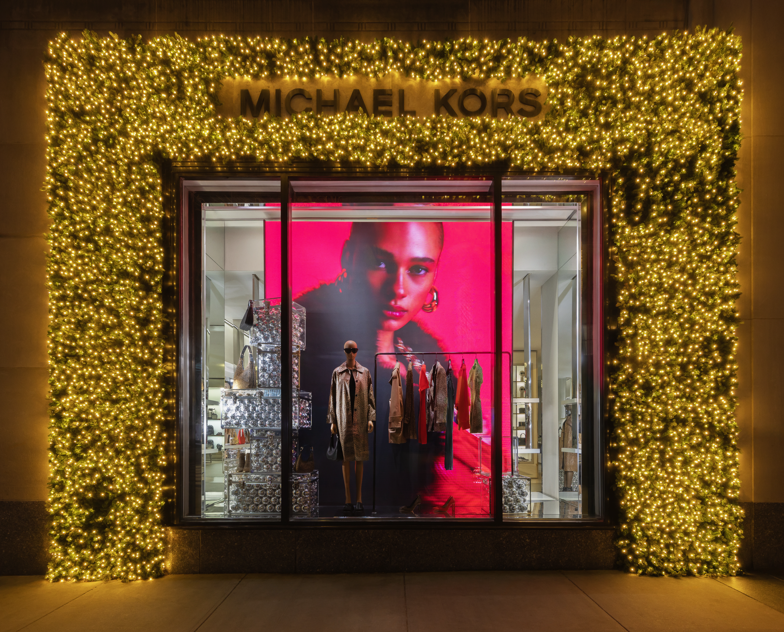 Storefront decorated with Christmas or holiday lights surrounding a Michael Kors store display window, featuring mannequins dressed in fashion clothing and accessories.