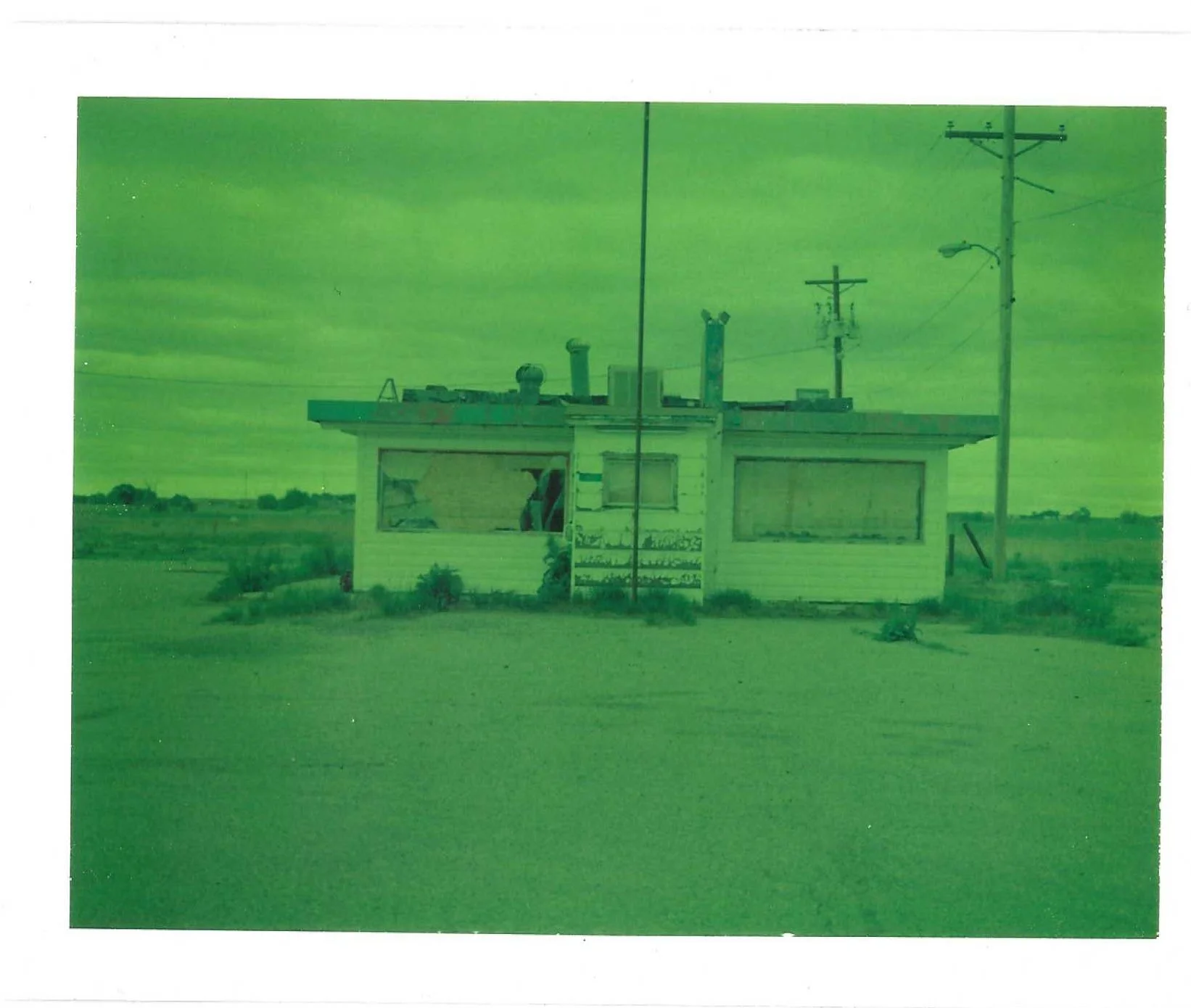 A small, weathered building with broken or boarded-up windows, rural surroundings, and electrical poles nearby, under overcast skies.