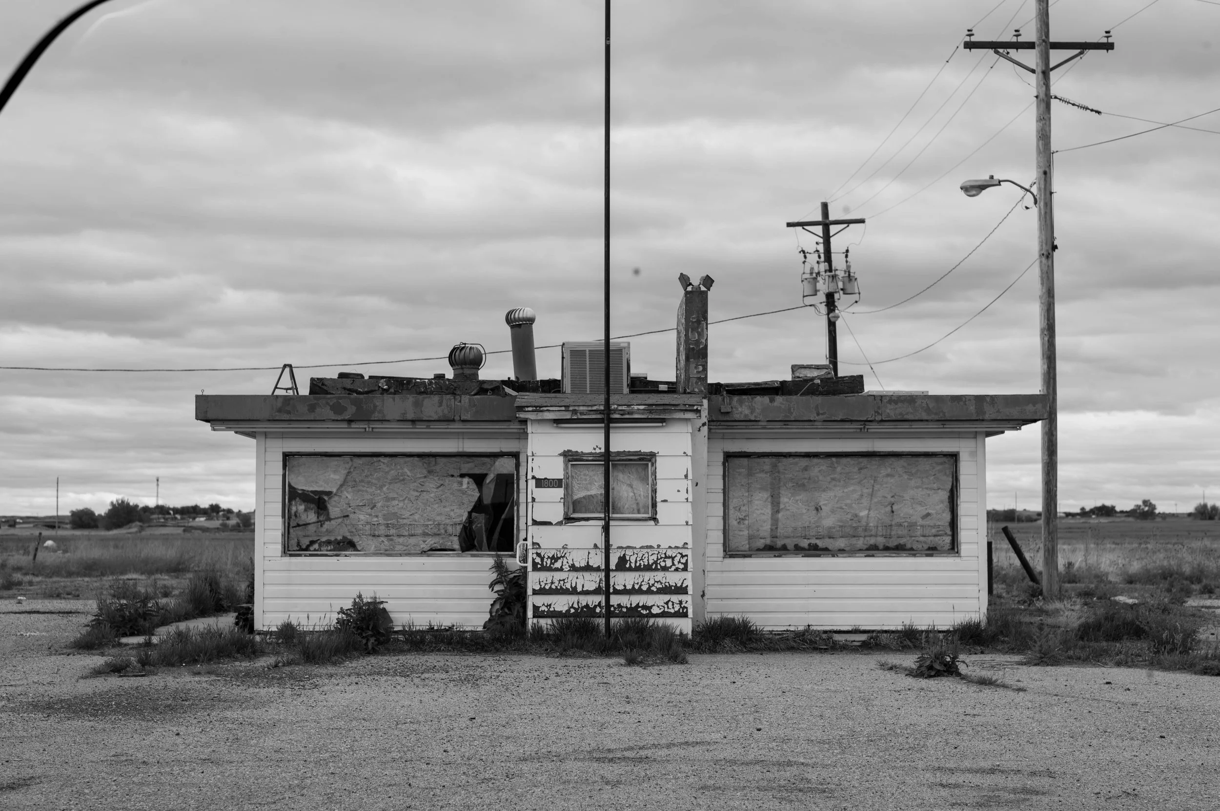 A weathered, abandoned building with boarded-up windows and peeling paint, situated in a rural area with open fields and overcast skies, featuring multiple utility poles and wires.