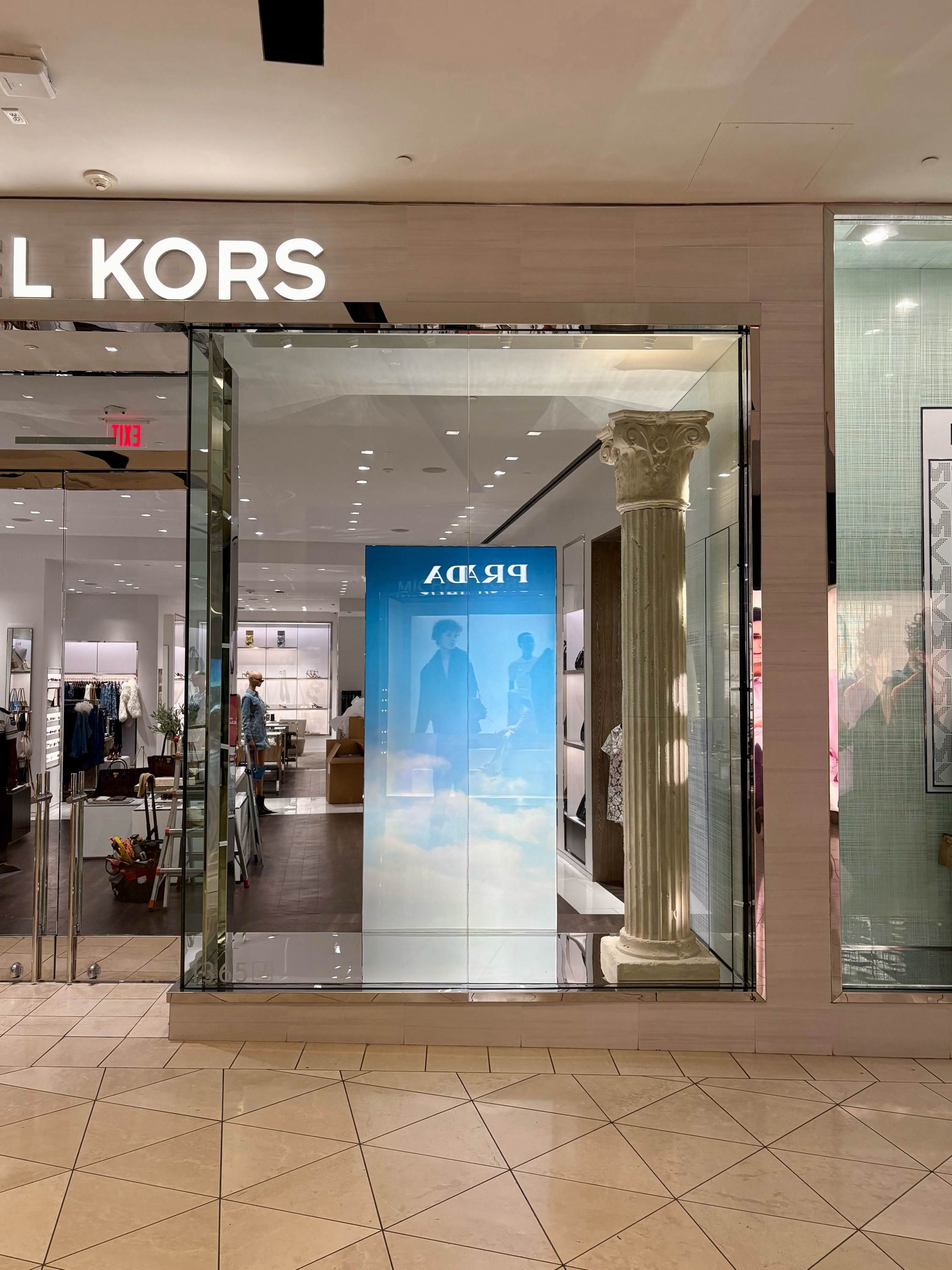 Entrance to a Michael Kors store in a shopping mall, with a glass window displaying a Roman-style column inside and clothing items visible in the background.