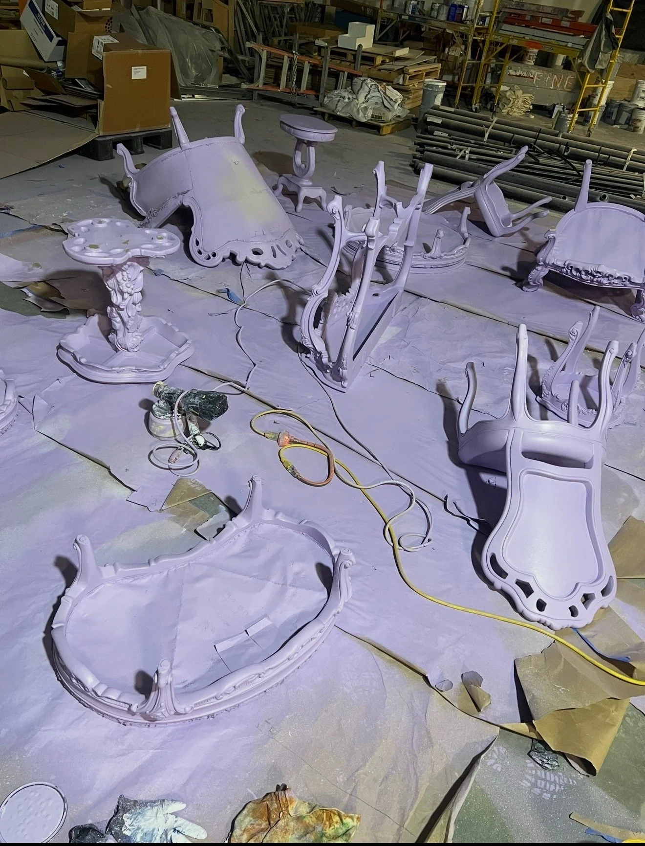 Furniture parts, including a table, chairs, and a cabinet, are seen in a workshop, with some pieces lying on the ground and others assembled.