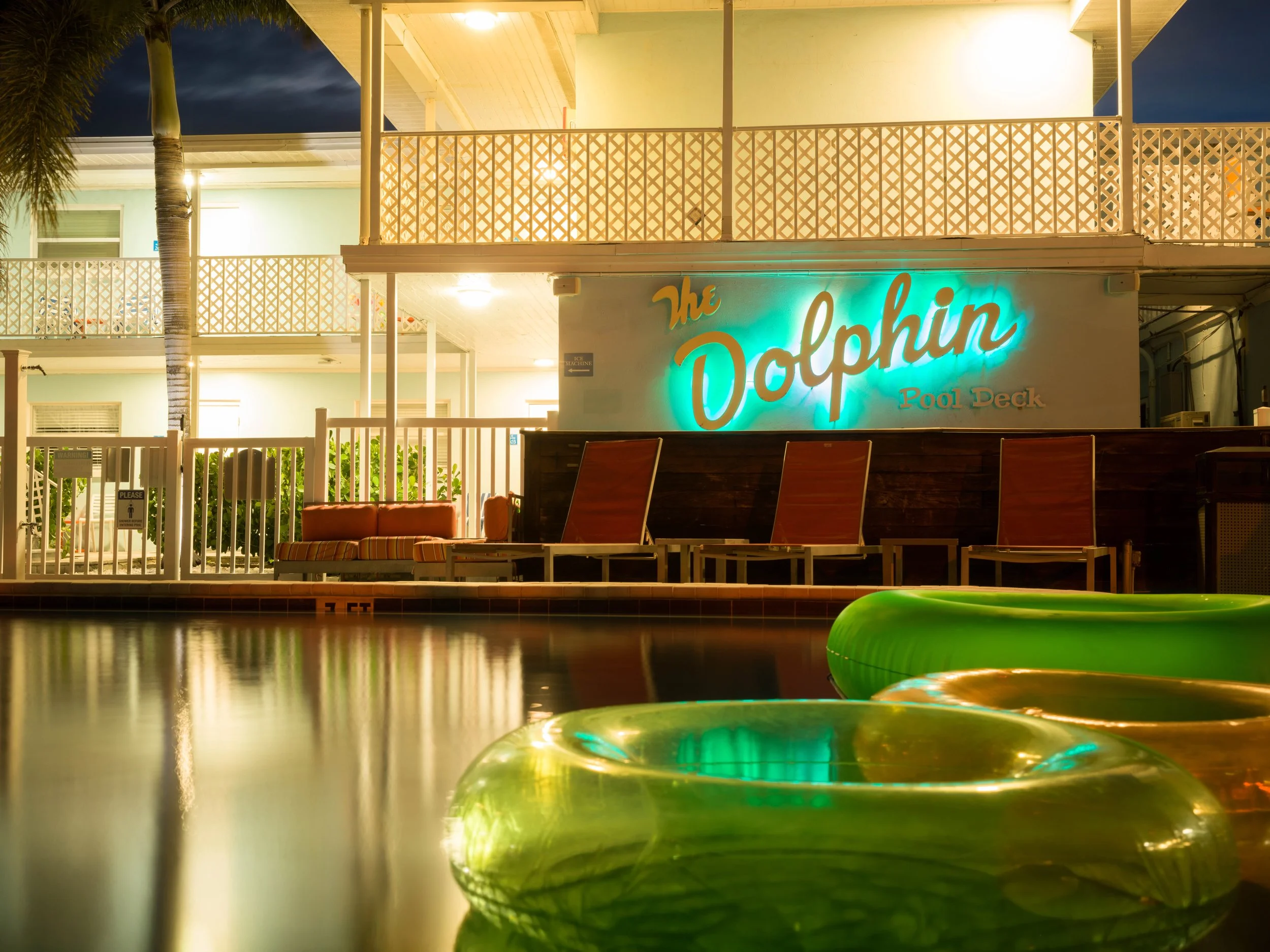 Night view of a poolside area with illuminated sign reading 'The Dolphin Pool Deck' and green pool floats reflecting in the water.
