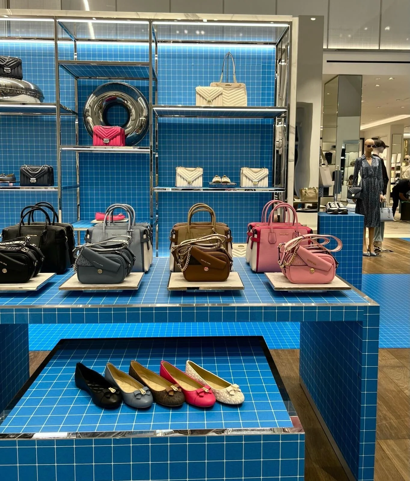 Display of handbags, wallets, and shoes on a blue-tiled table and shelves in a retail store.
