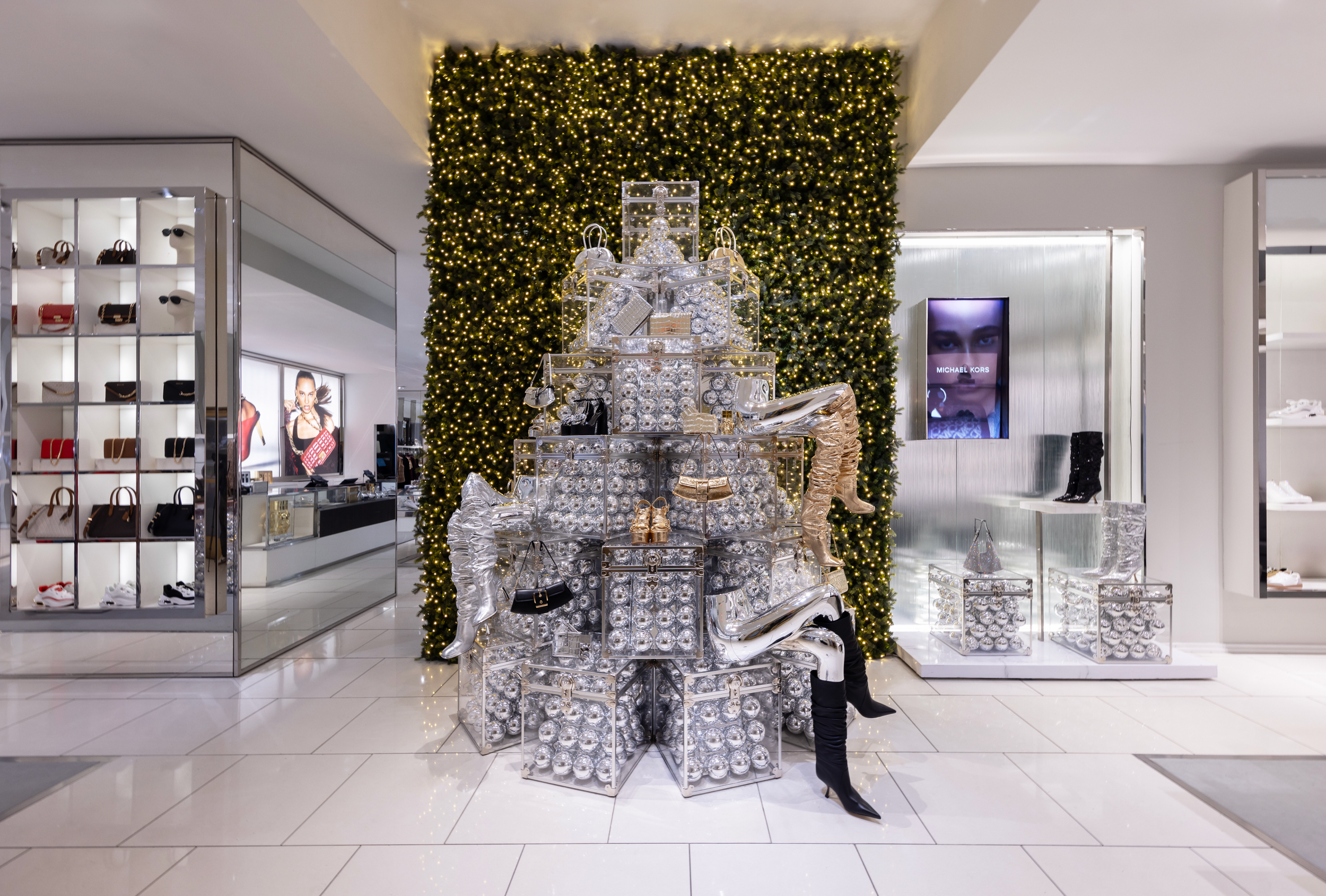 Decorative display in a shopping mall featuring a large green wall of Christmas lights, with a pyramid-shaped arrangement of silver ornaments and festive accessories such as boots, handbags, and shoes, with store signage and advertisements in the bac