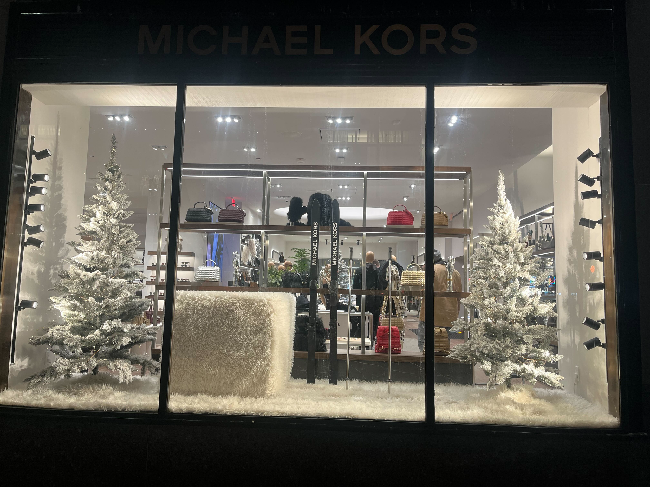 Front view of a Michael Kors store window display with two white Christmas trees, handbags, and mannequins inside.