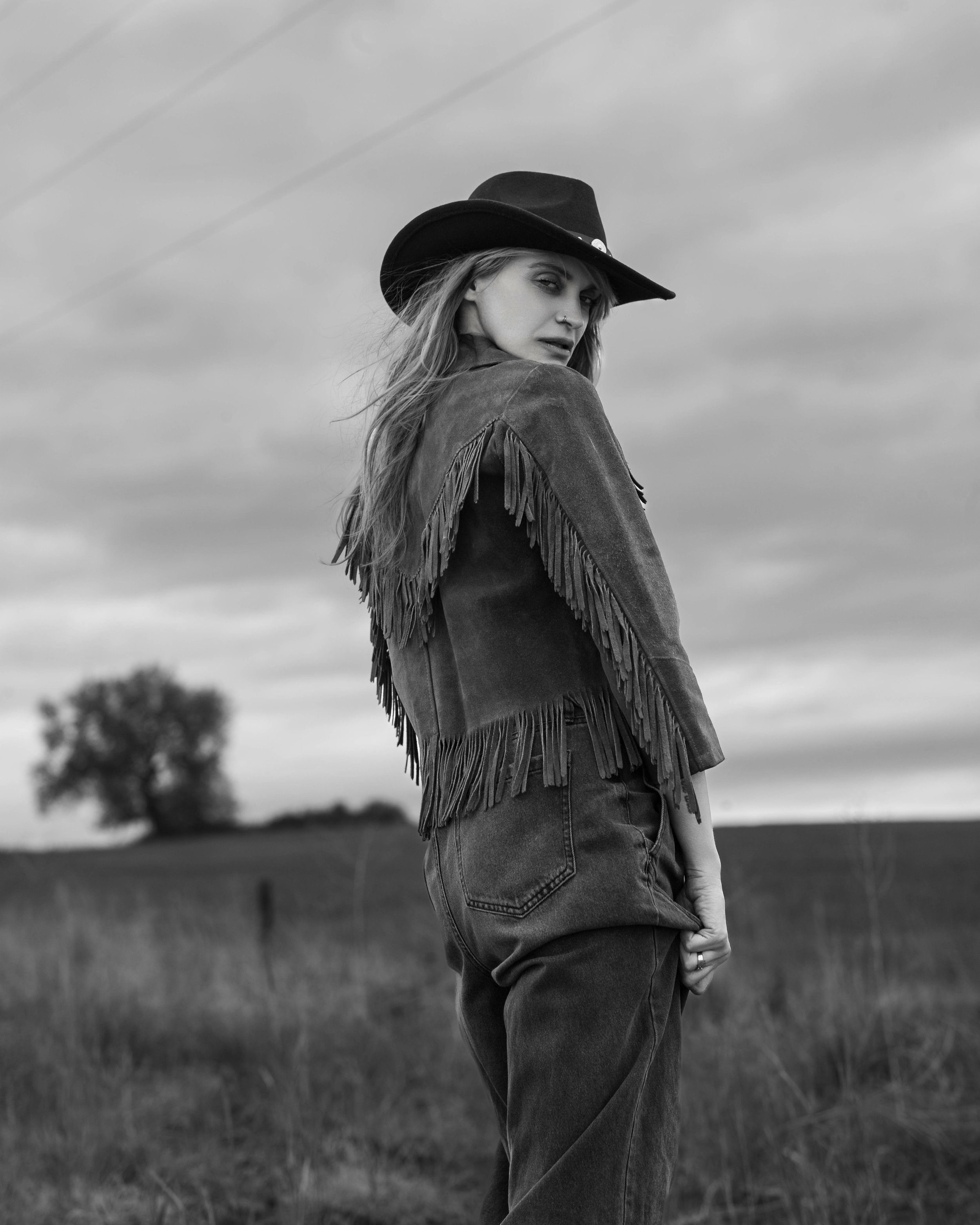 A woman in a wide-brimmed hat and fringed jacket standing outdoors in a field with a tree in the distance, captured in black and white with cloudy sky.