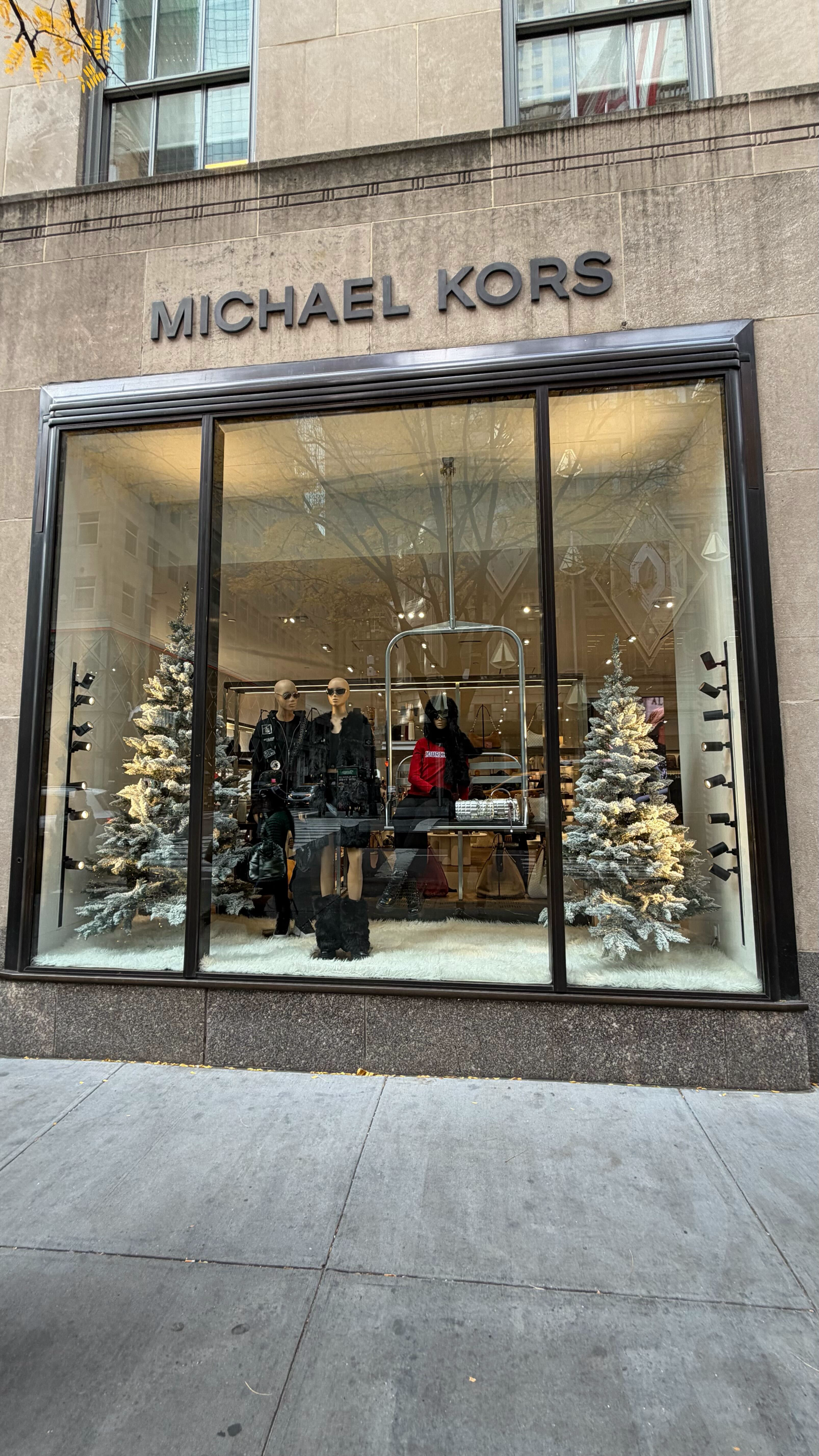 Storefront window display for Michael Kors with mannequins dressed in clothing, surrounded by two decorated snow-covered Christmas trees.