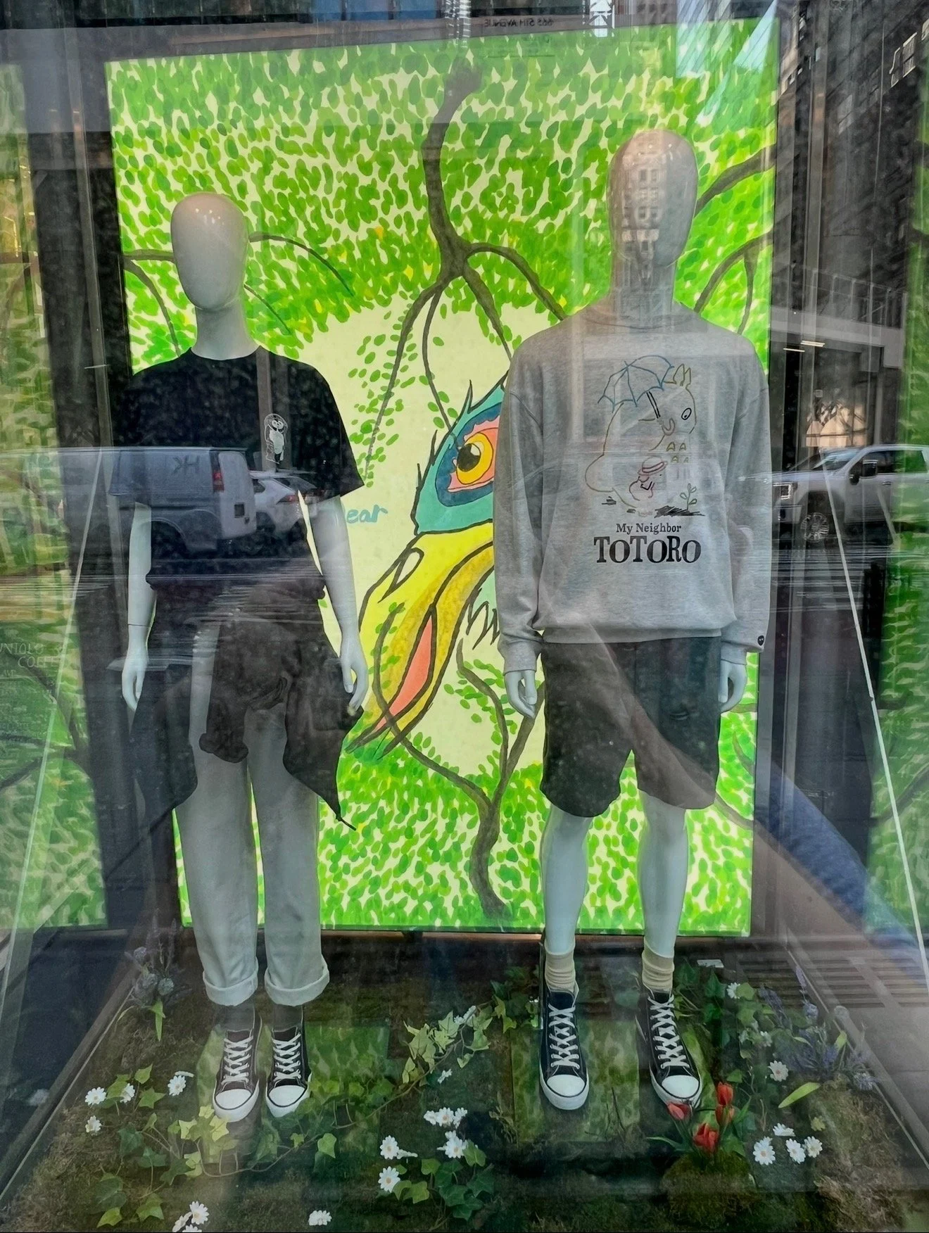 Mannequins dressed in casual clothing displayed in a store window, with a colorful illustration of a bird and plant background behind them, and a glass reflection of the street outside.