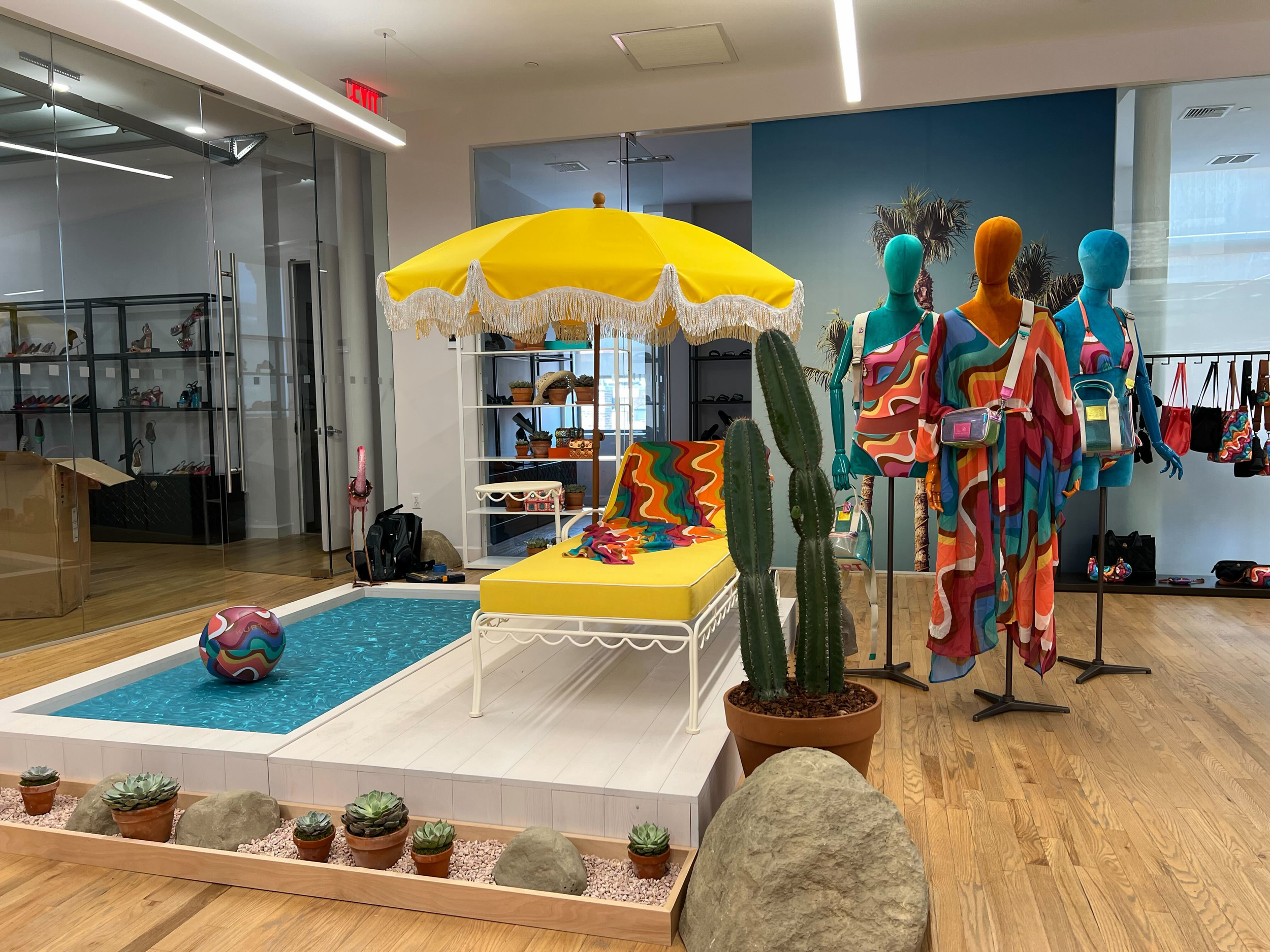 Indoor retail display with mannequins dressed in colorful patterned clothing, large cactus plants, an yellow umbrella with fringe, and a small pool with a colorful ball, surrounded by succulents and large rocks.