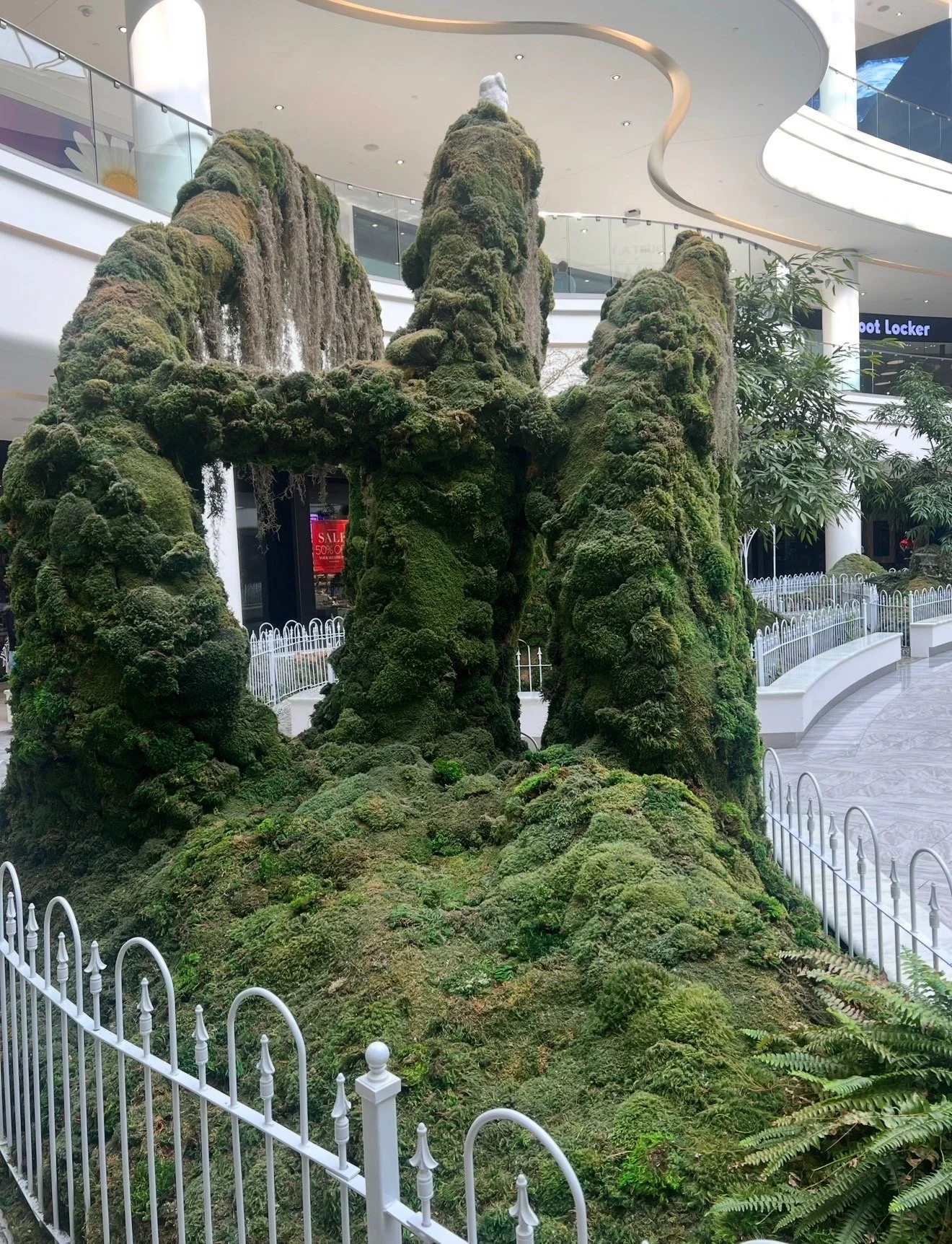 Indoor moss sculpture resembling a mountain landscape with a waterfall, surrounded by white fence and located in shopping mall with modern architecture and stores visible in background.