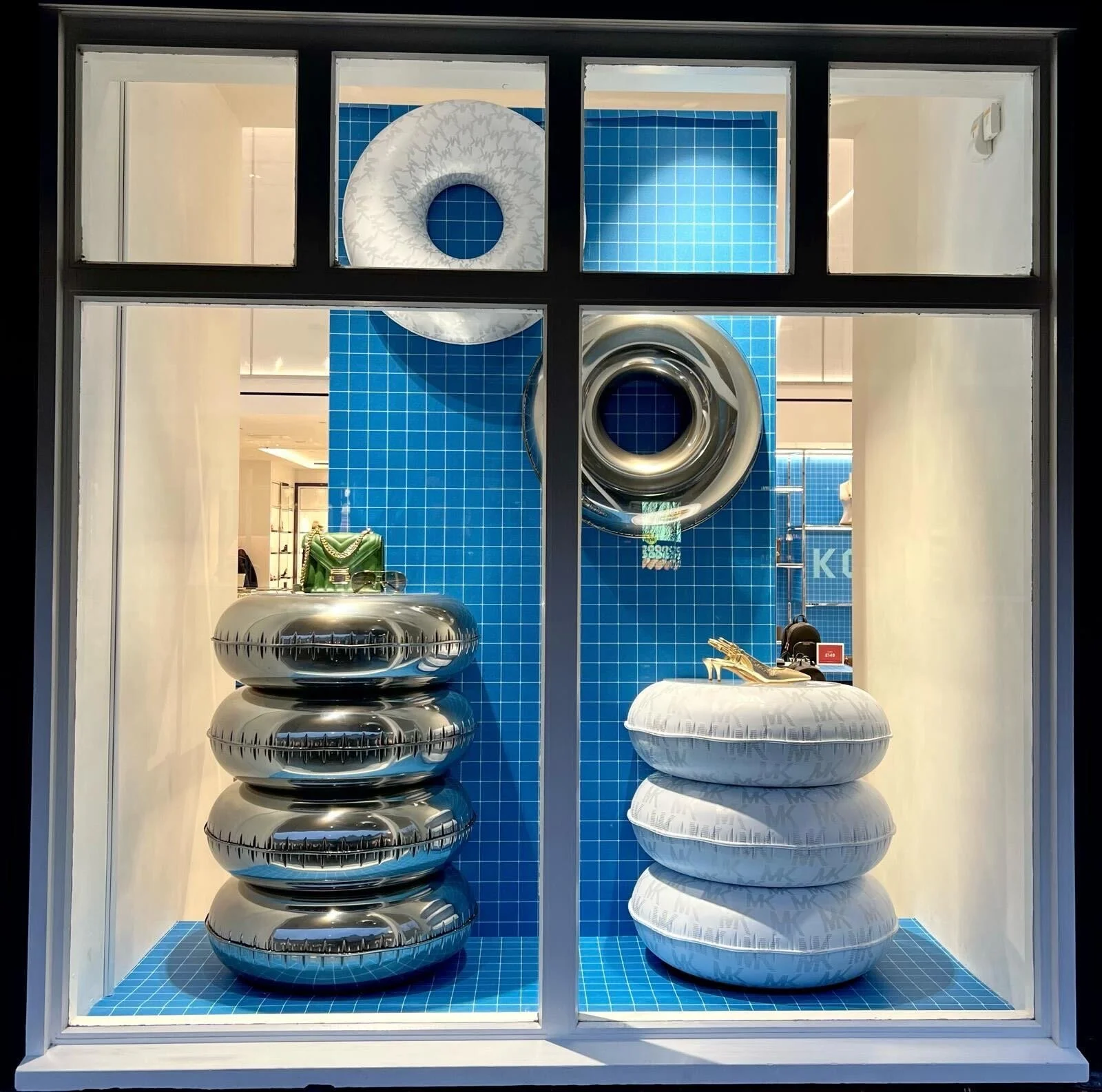 Display window showcasing metallic and white pillows, blue-tiled wall background, and decorative items including a green handbag and gold shoes.