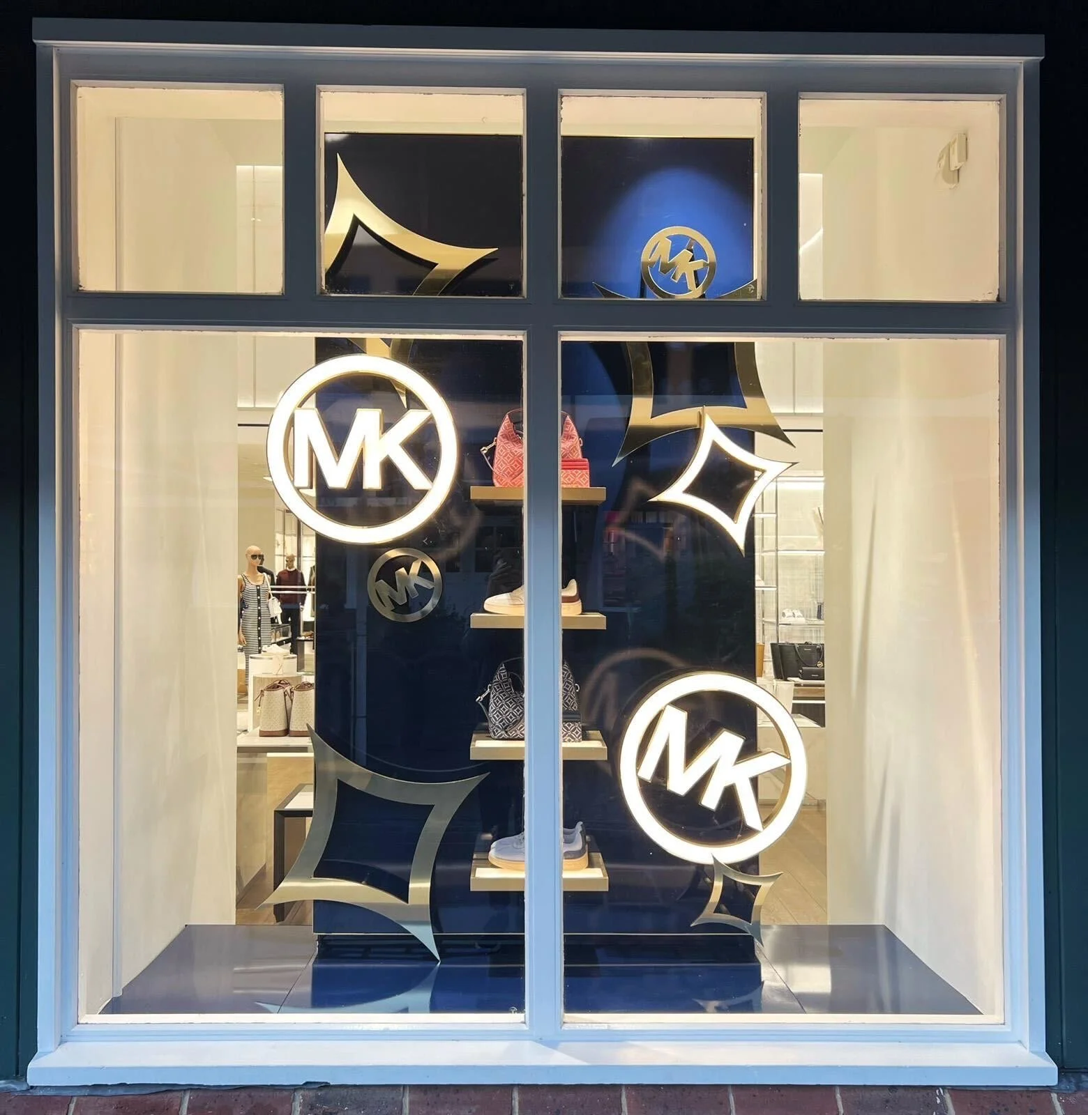 Store window displaying Michael Kors handbags and accessories, with prominent MK logo signs and decorative black and gold designs, seen through glass.