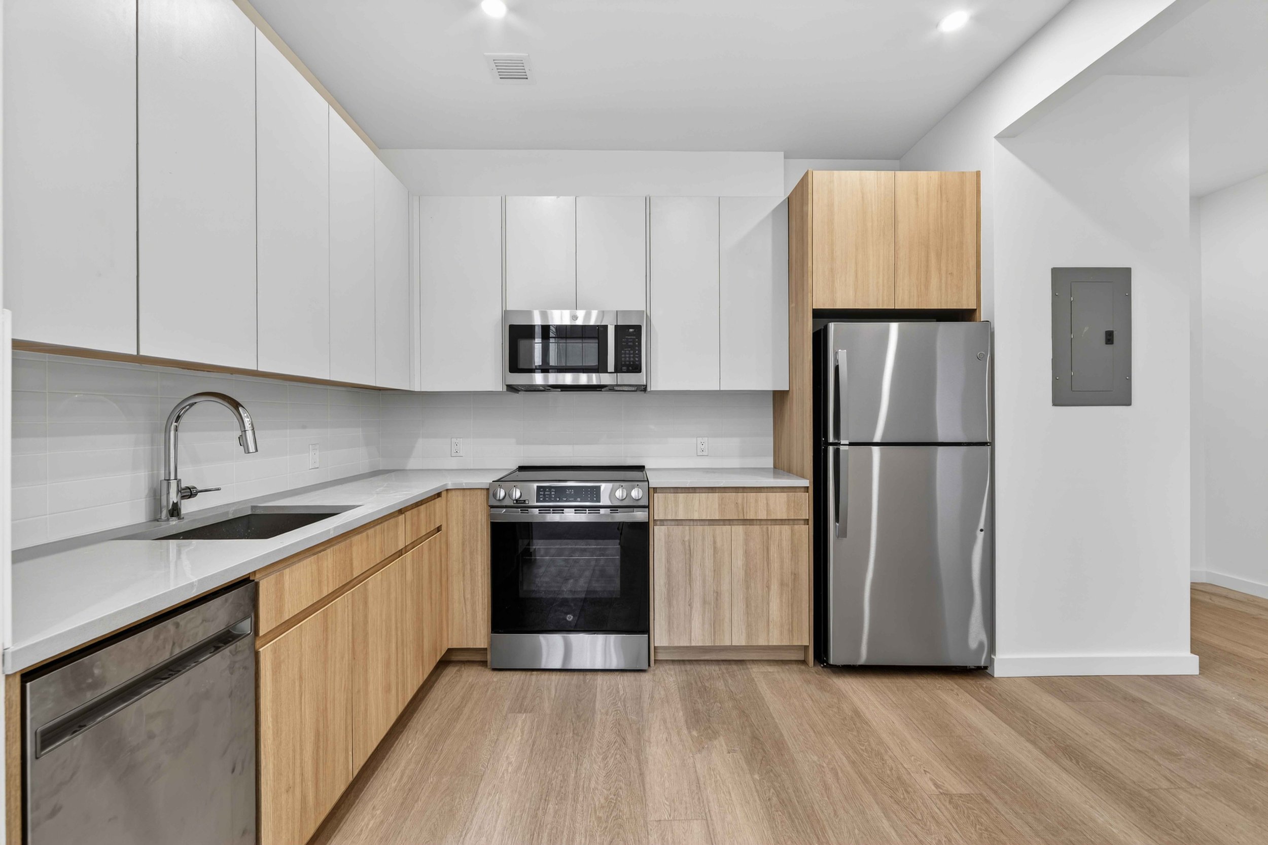 A modern kitchen with white upper cabinets, wooden lower cabinets, stainless steel appliances including a microwave, oven, and refrigerator, and a white countertop with a sink and faucet.