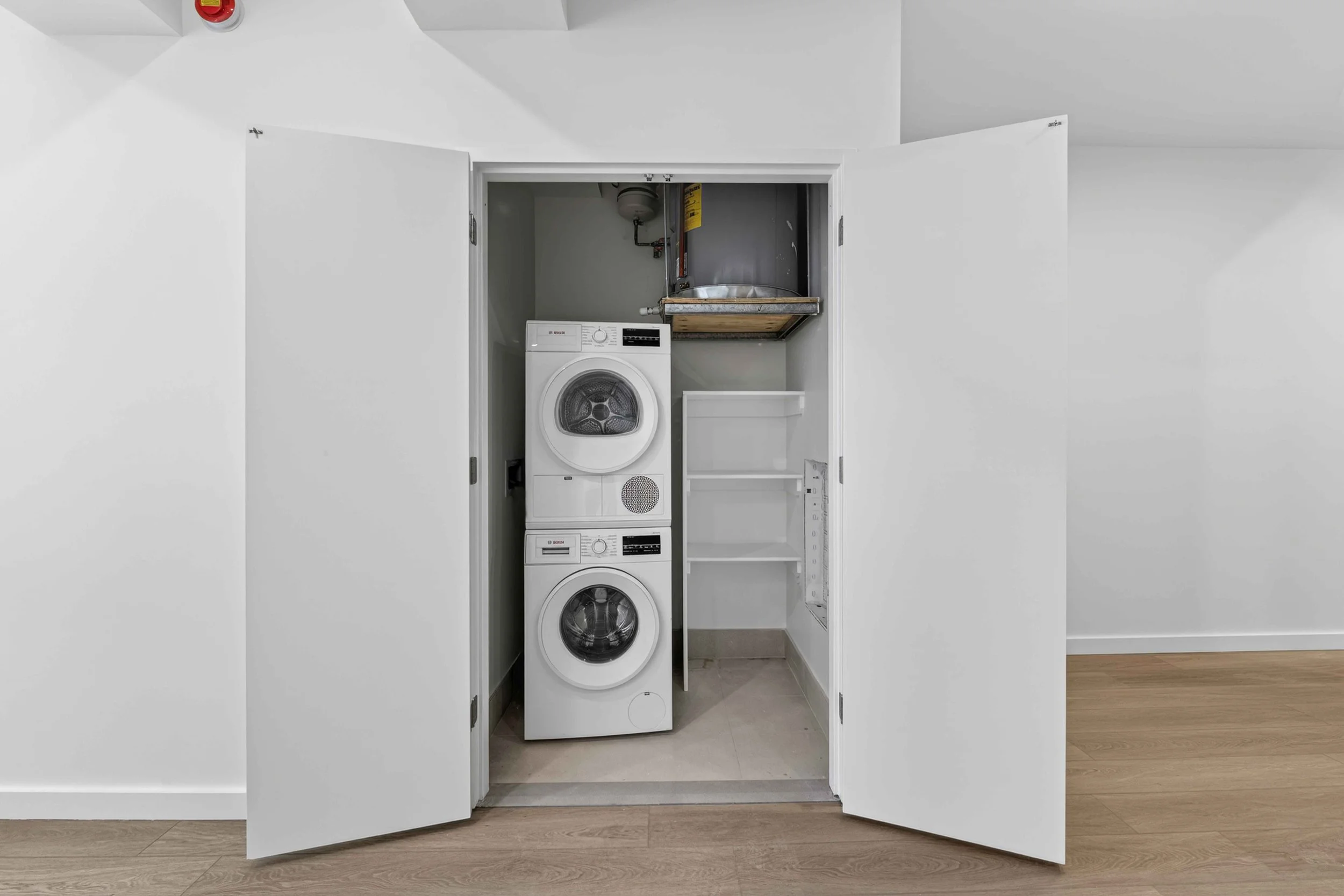 A laundry closet with white double doors open, revealing stacked washer and dryer, a sink, and empty shelving.