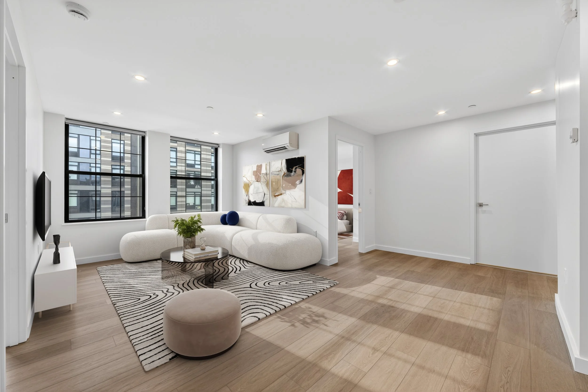 Modern living room with large windows, white walls, a white sofa, a round wooden coffee table, abstract wall art, a patterned rug, and wooden flooring.