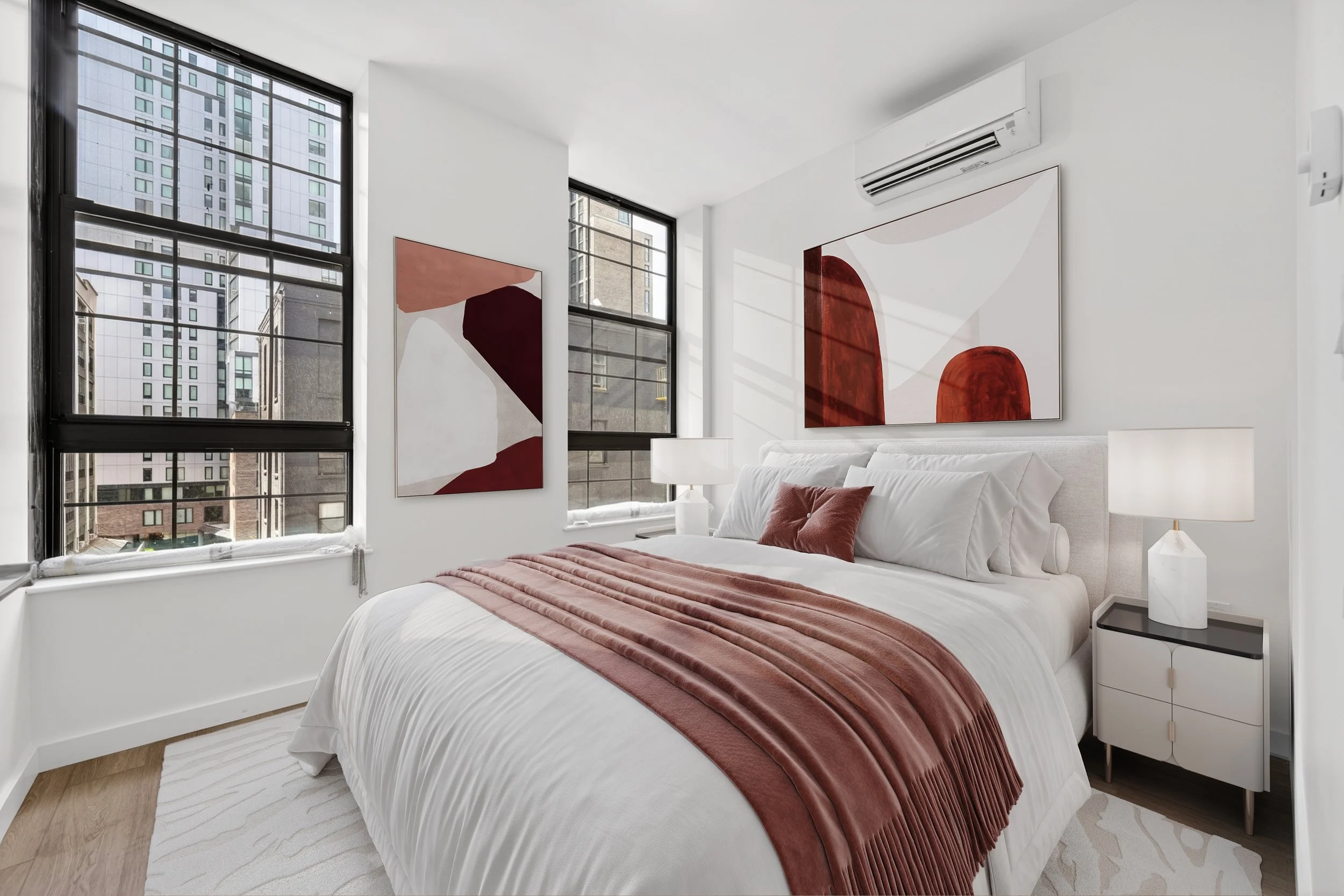 Modern bedroom with white walls, large windows showing city buildings, abstract wall art, white bedding, rust-colored throw, and bedside table with lamp.