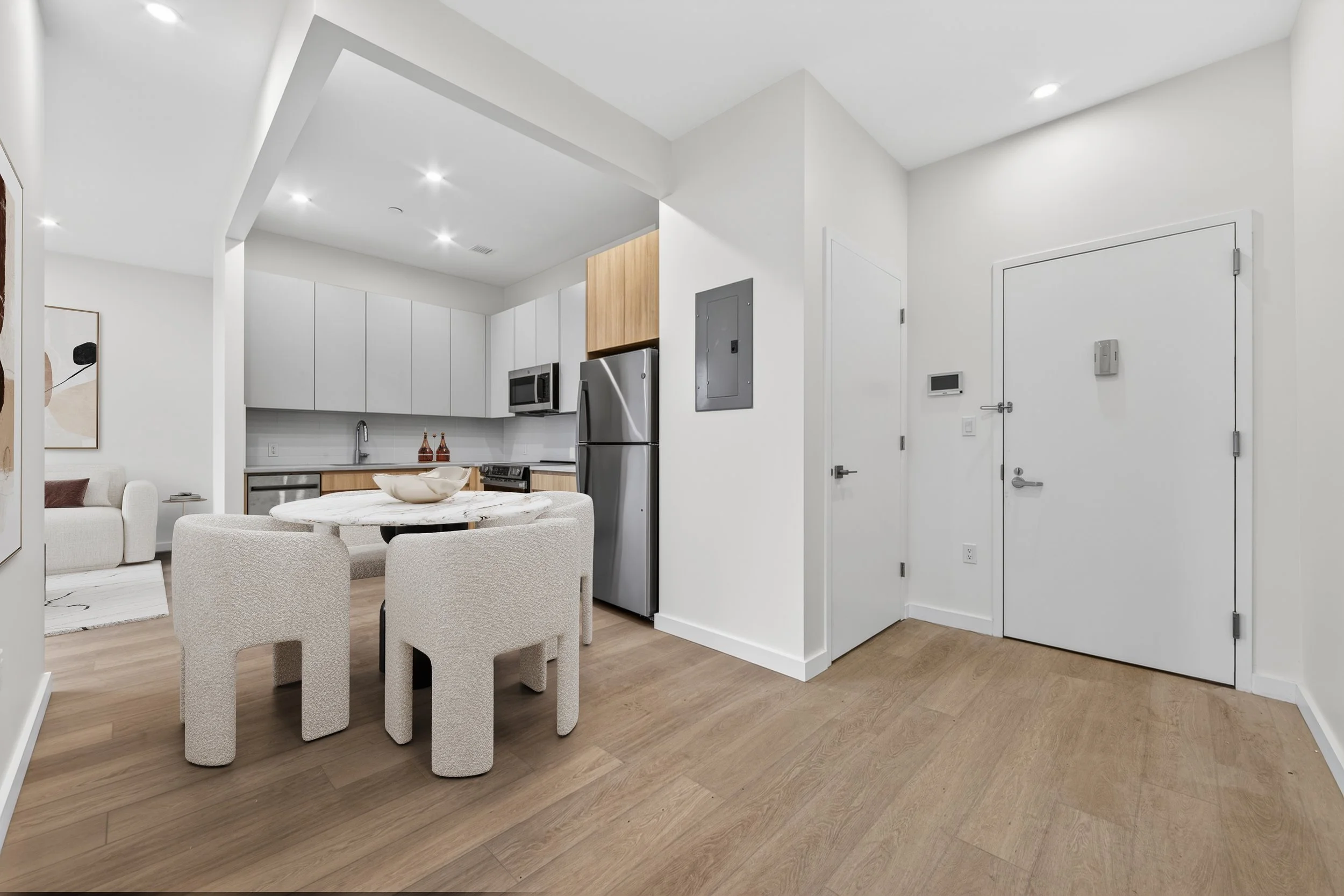 Modern kitchen and dining area with white cabinets, stainless steel refrigerator, microwave, and a round marble table with four beige textured chairs. There is a living room with a white sofa and abstract artwork on the wall. The flooring is light wo
