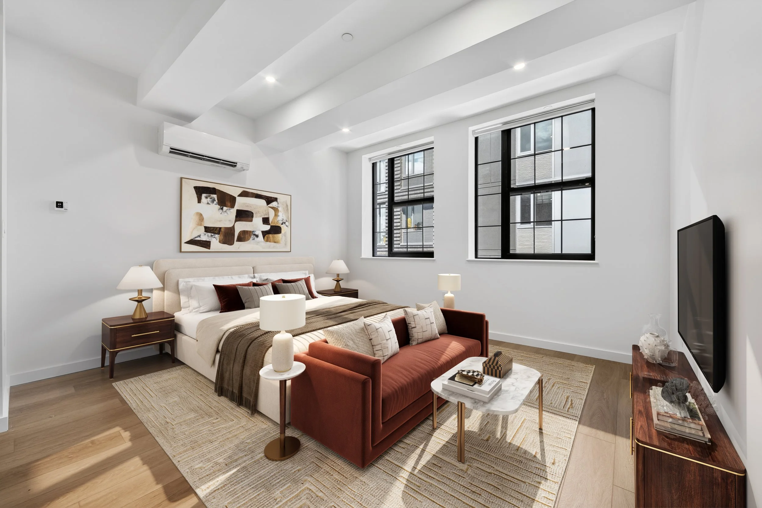 Modern bedroom with white walls, a bed with beige and rust-colored pillows, two nightstands with lamps, large windows with black frames, abstract wall art, a rust-colored sofa, a small marble coffee table with books, and a television on a wooden stan