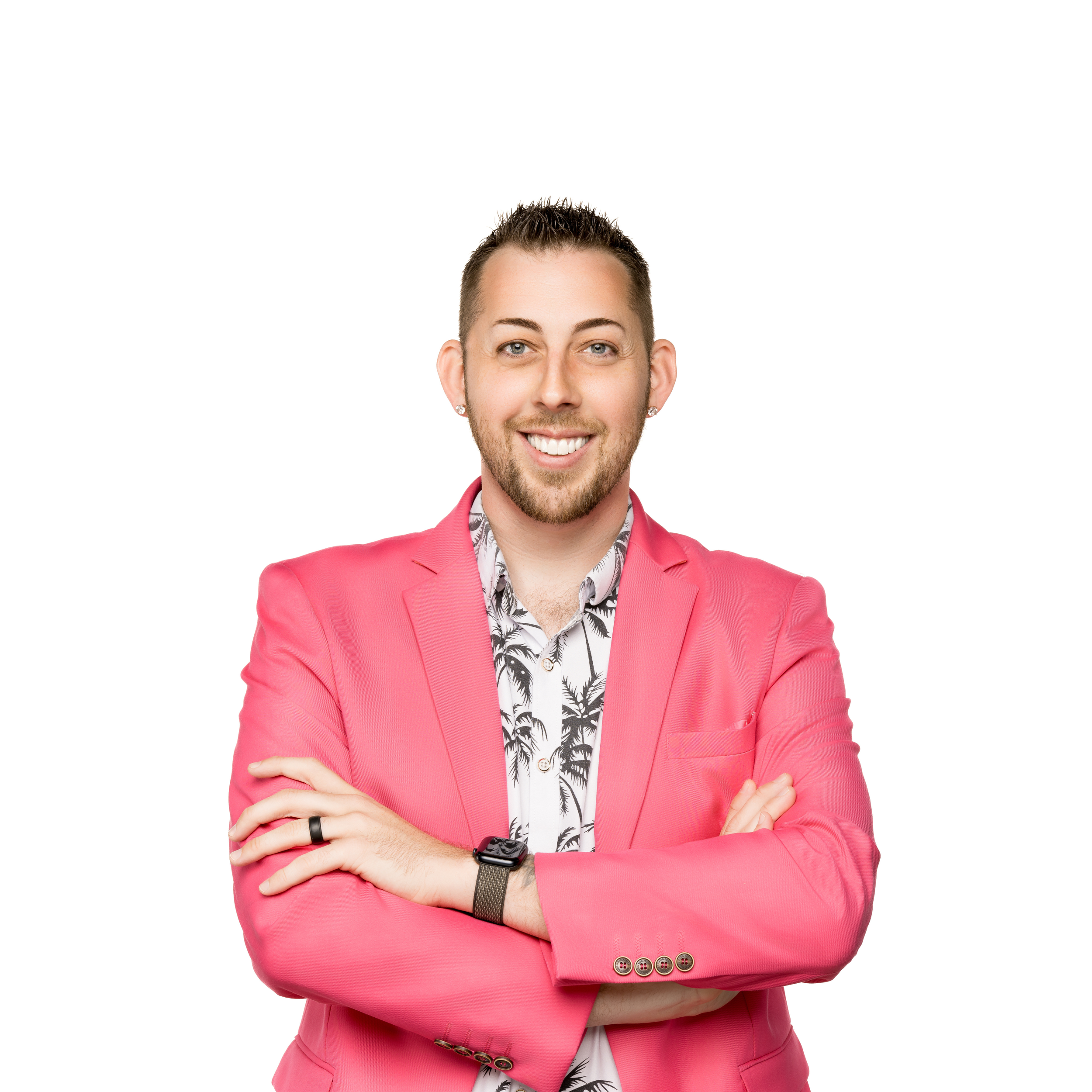 A man with short spiked hair, light eyes, a beard, and earrings is smiling, wearing a pink blazer, a white shirt with black palm tree patterns, a black watch, and a black ring on his right hand, against a white background.