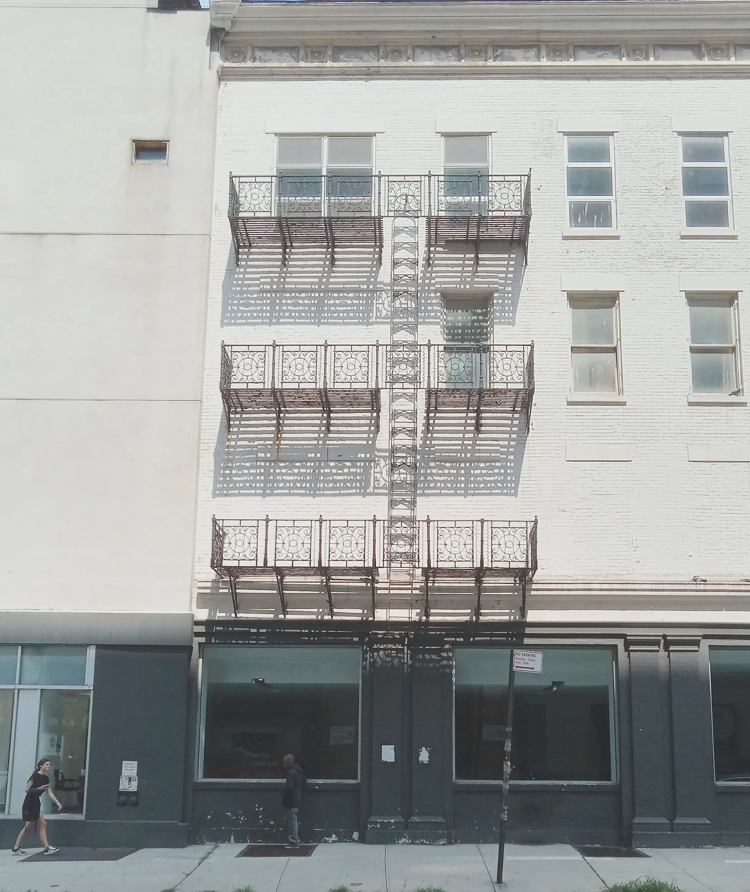 A white brick building with ornate fire escape balconies and glass storefront windows below. Two people are walking on the sidewalk in front of the building. One is a woman wearing a black outfit, and the other is a man in darker clothing.