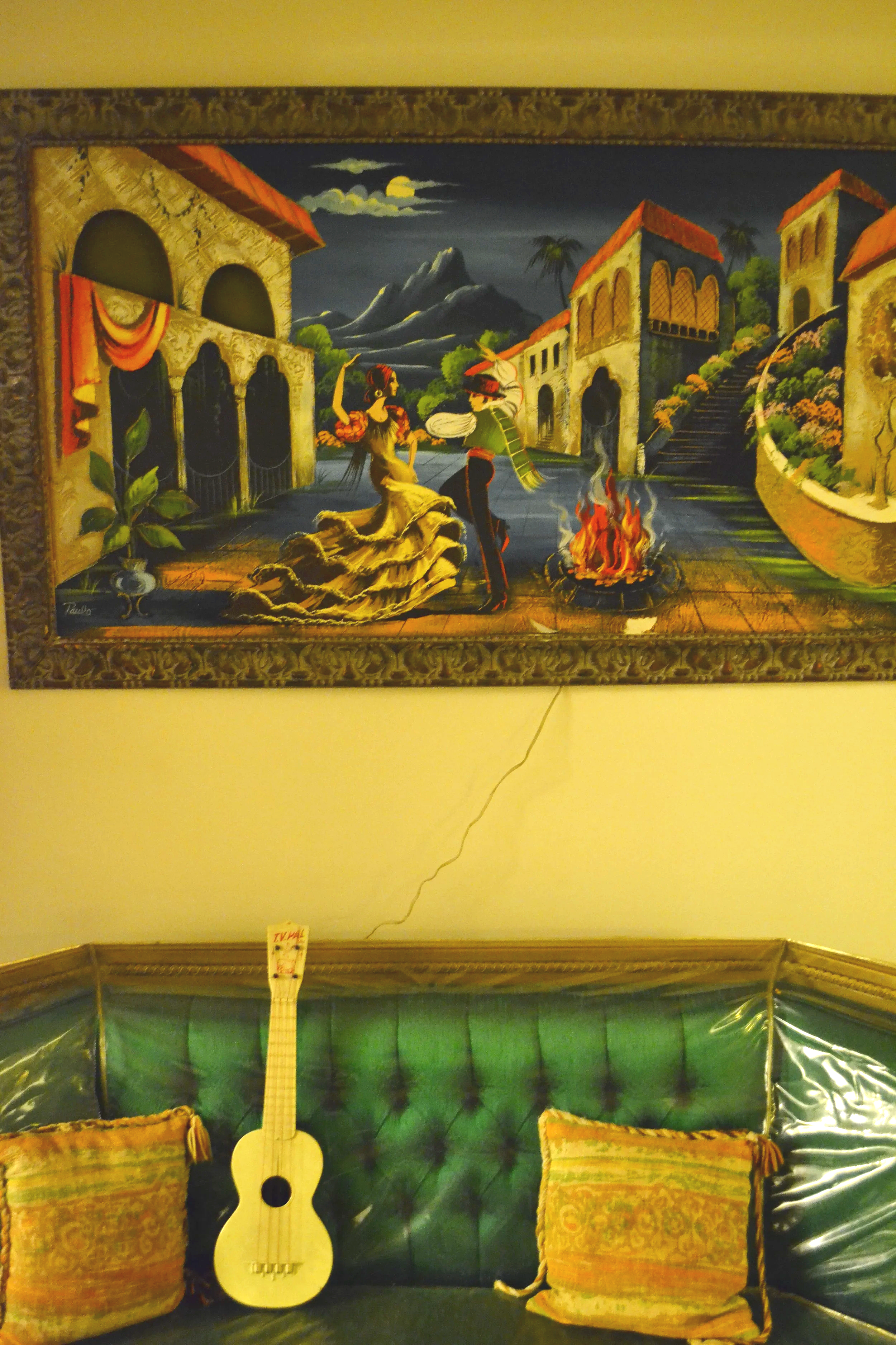 A mural depicting a scene with a woman in a yellow dress dancing near a campfire, a man playing a guitar, and traditional buildings under a moonlit sky with mountains in the background. There are pillows in the scene.