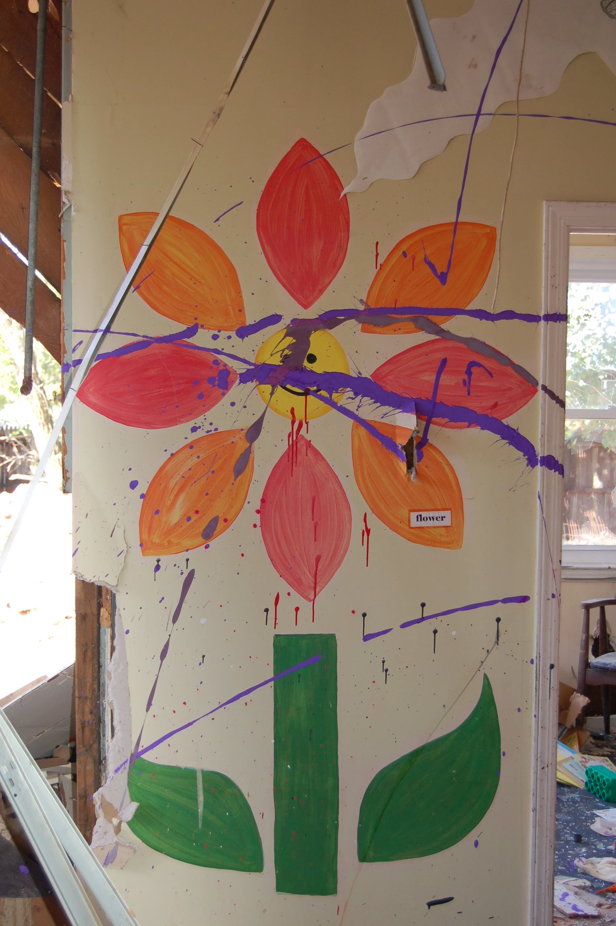 A colorful painted flower on a wall with large orange and red petals, a yellow center, green leaves, and purple splashes of paint in the background.