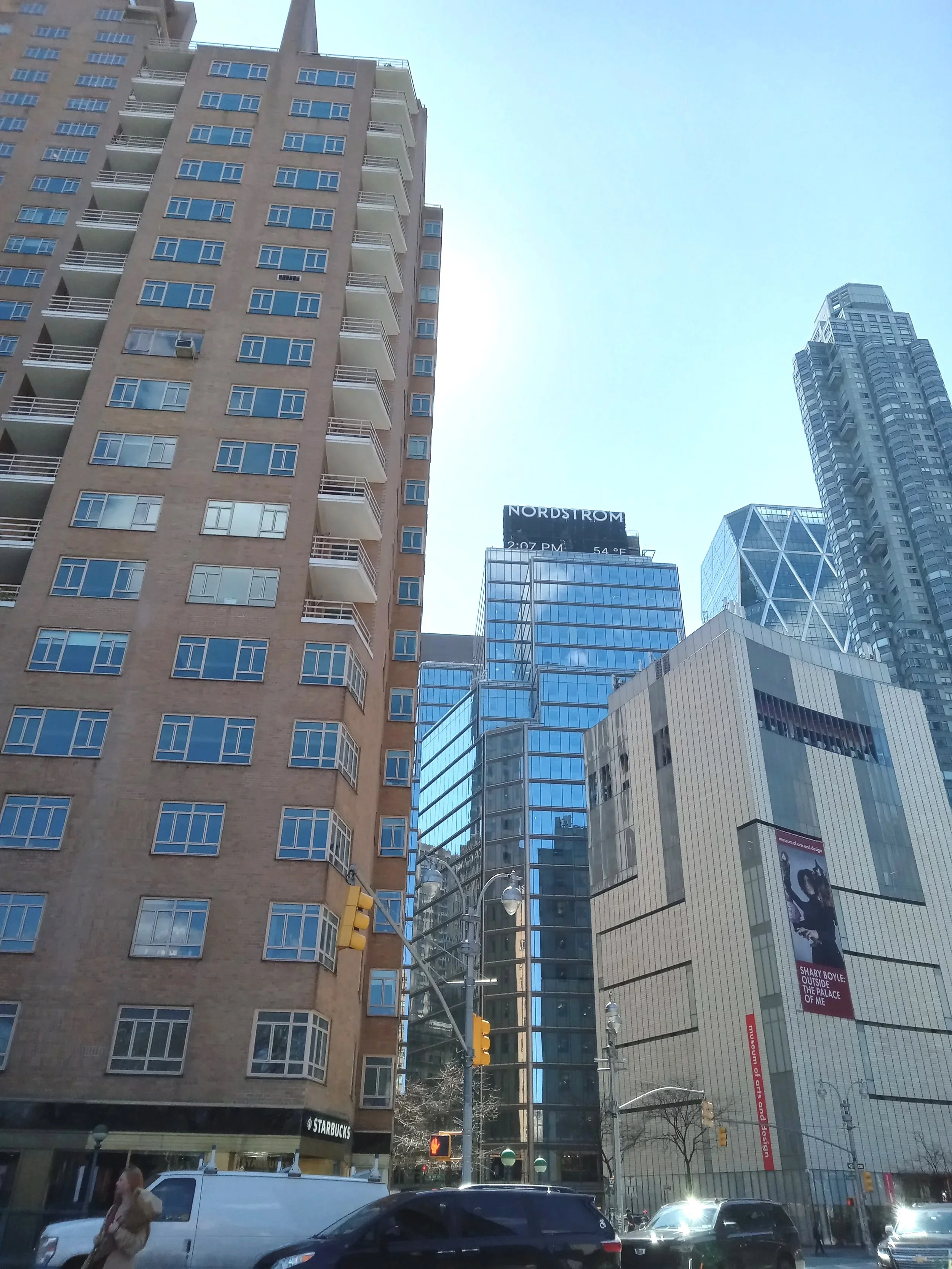 City street scene with tall buildings, including a beige apartment building on the left, a glass skyscraper in the middle, and a modern building with a museum sign on the right. There are cars and pedestrians on the street, and a digital billboard and traffic lights are visible.