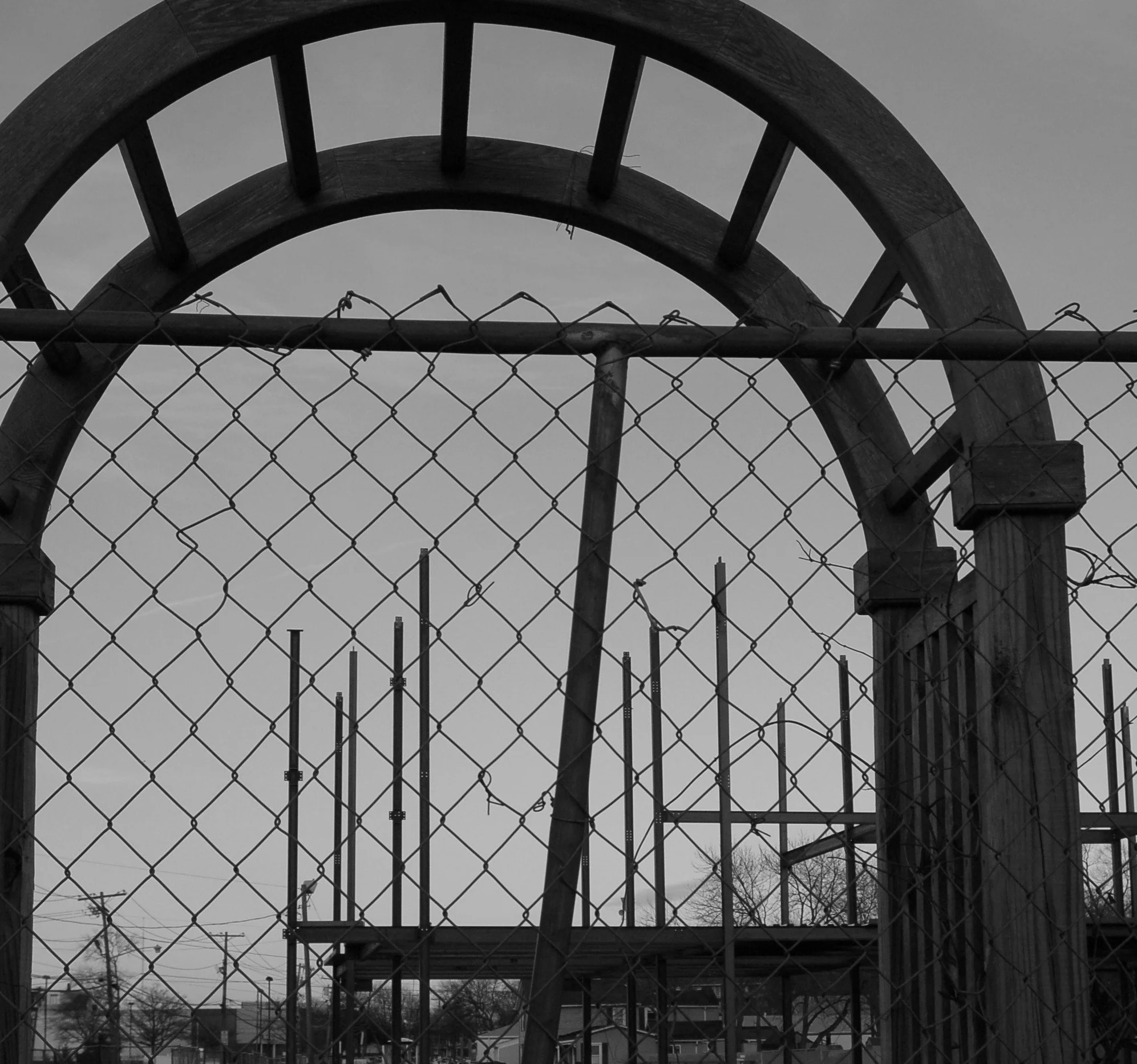 Black and white photo with a chain-link fence and tree branches and buildings in the background under an overcast sky.