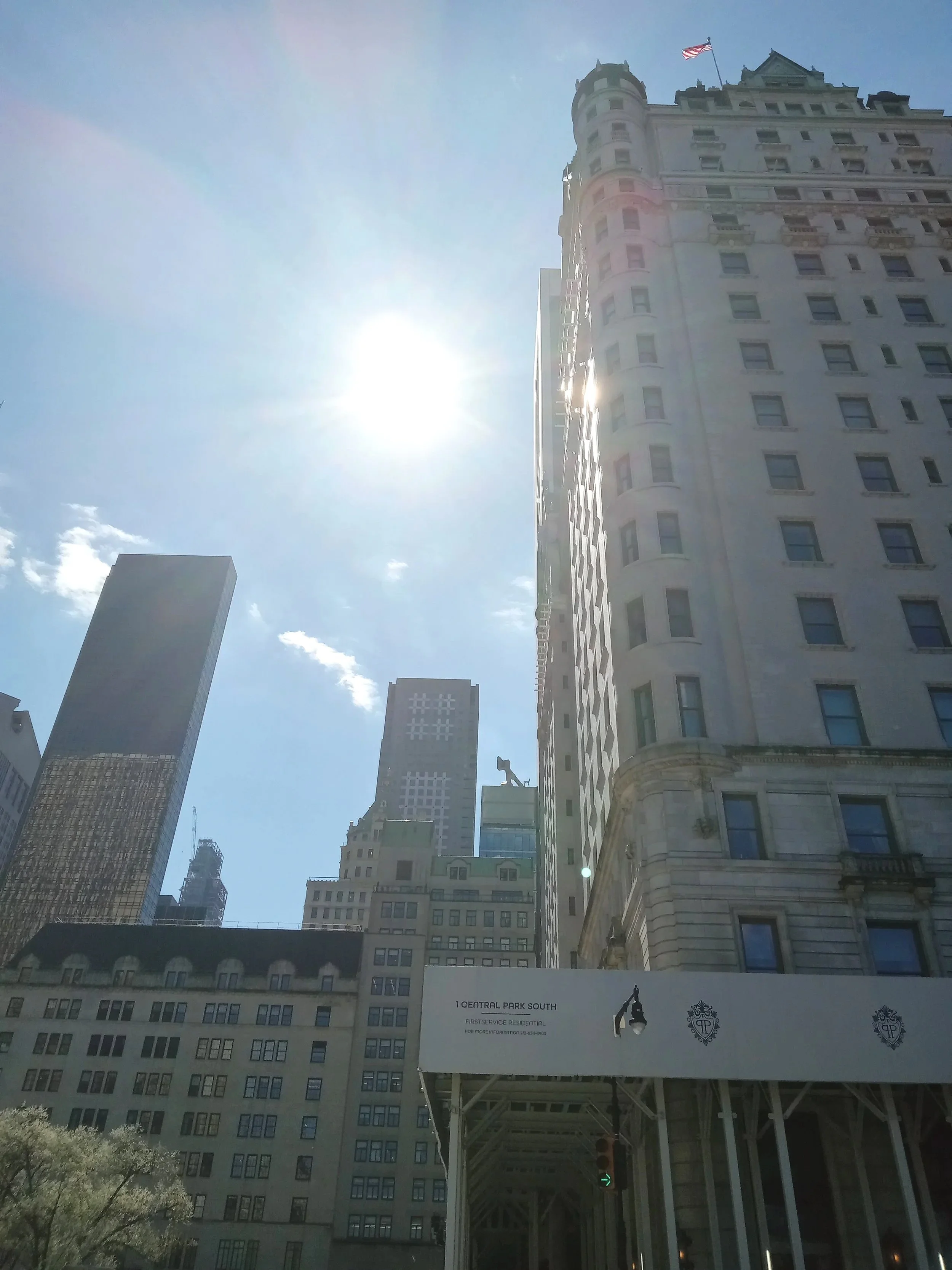 Skyscrapers in an urban cityscape with the sun shining brightly in a clear blue sky, American flags flying on top of some buildings, and road signs visible at the street level.