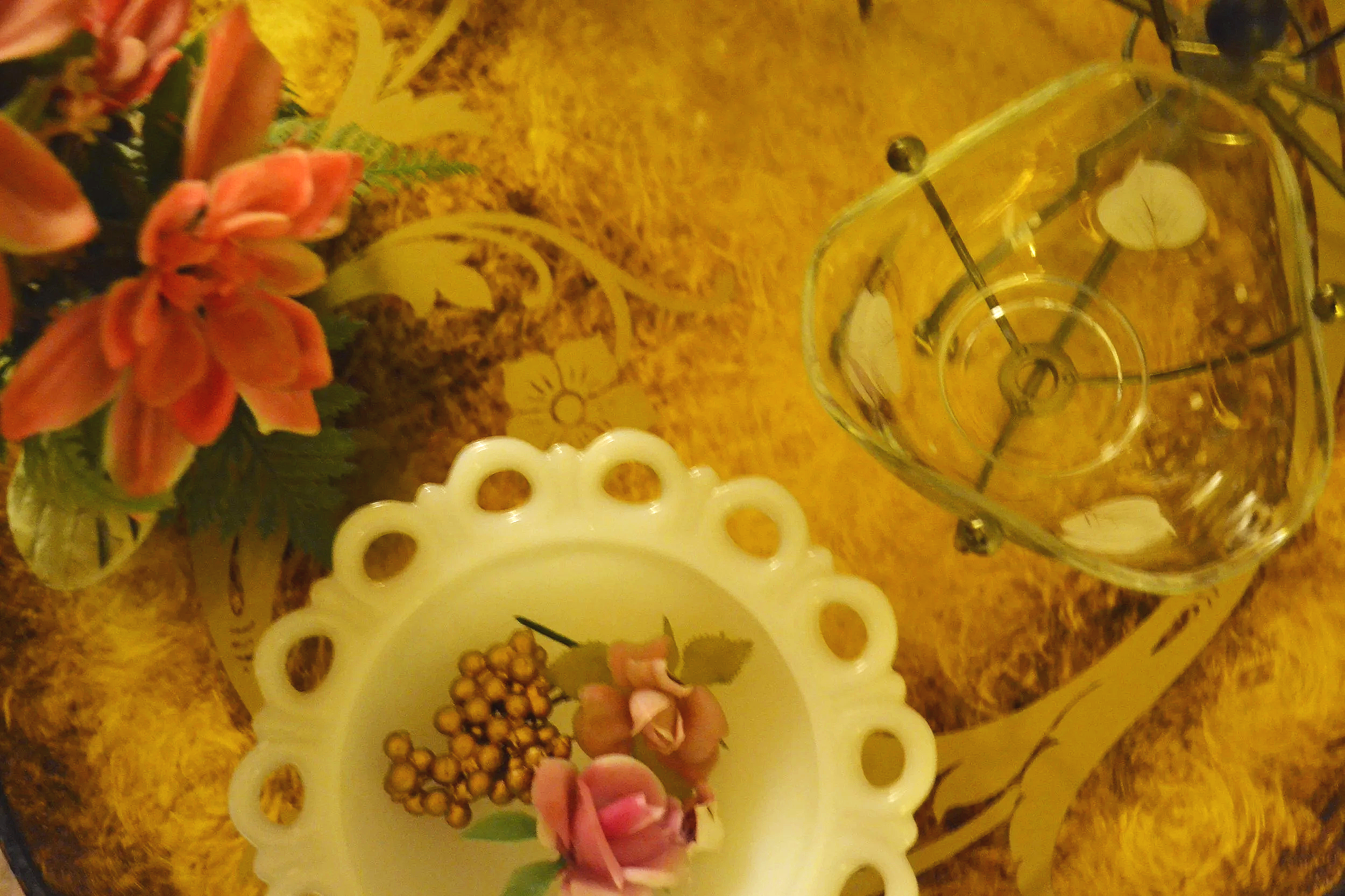 A glass vase on a table holding orange and pink floral arrangements, a decorative white plate with cut-out patterns, a small dish with pink petals and a cluster of small round objects, all on a gold patterned table.