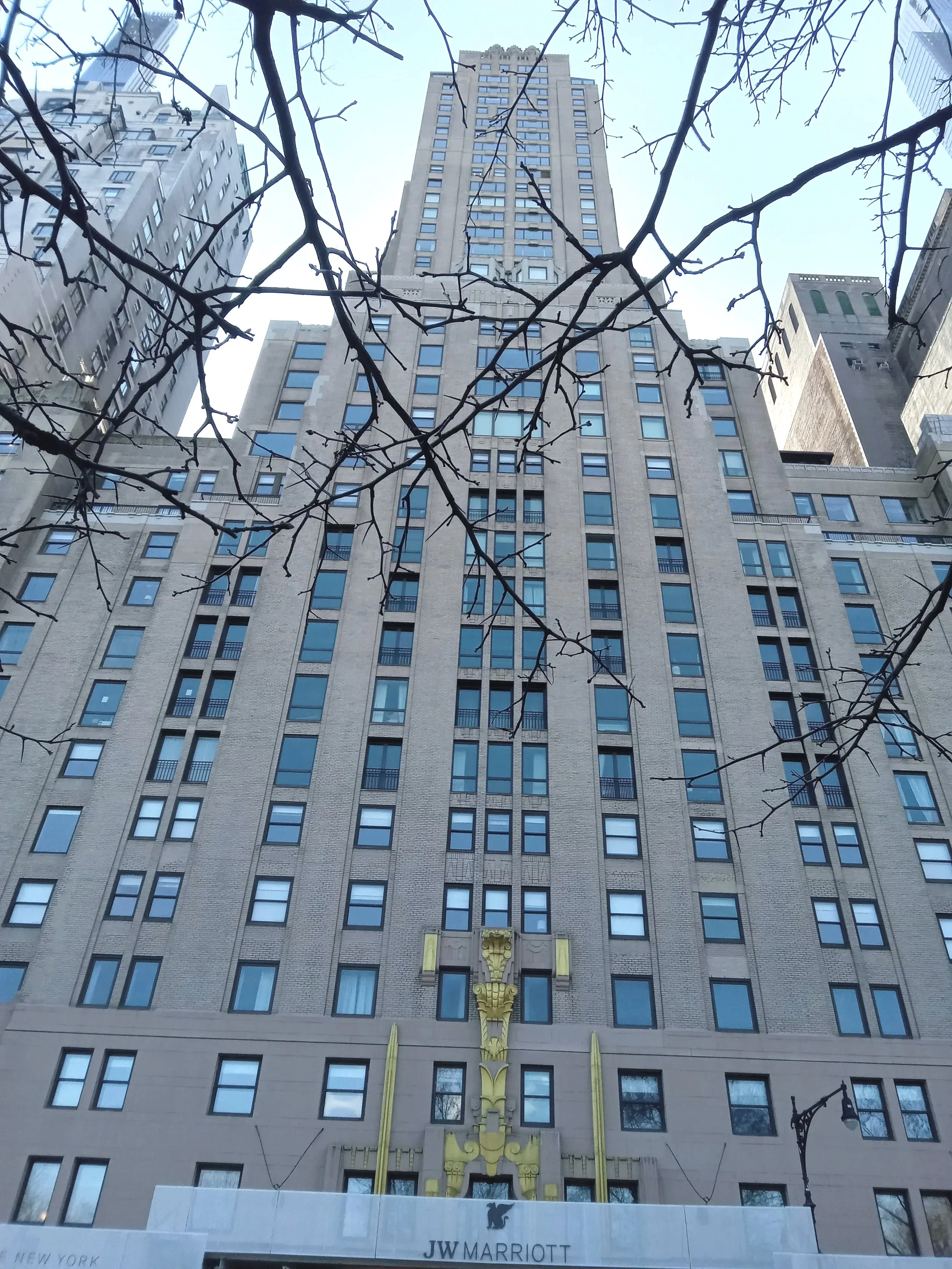 Tall hotel building with the JW Marriott logo at the base, decorative gold architectural elements on the facade, and leafless tree branches in the foreground in New York.