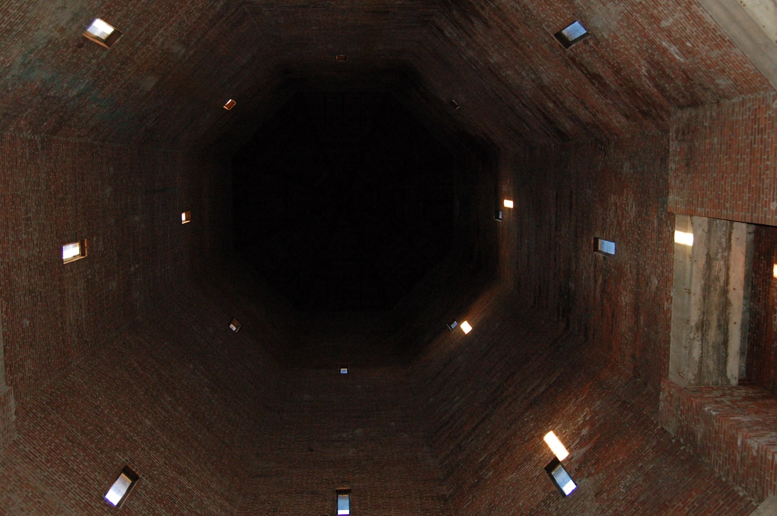 Looking up at the interior of a large brick dome or tower with small rectangular windows emitting light.