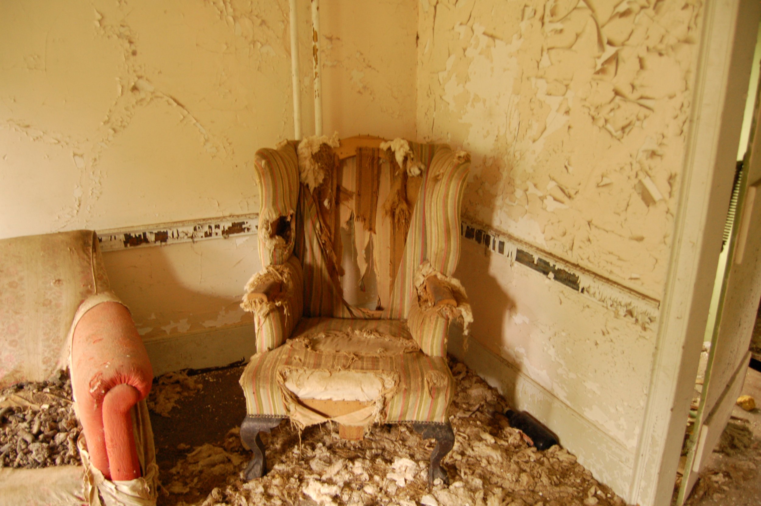 A room with damaged furniture and peeling paint on the walls. There is an old, torn armchair with multicolored striped upholstery, a discolored and tattered sofa, and debris on the floor, indicating renovation or destruction.