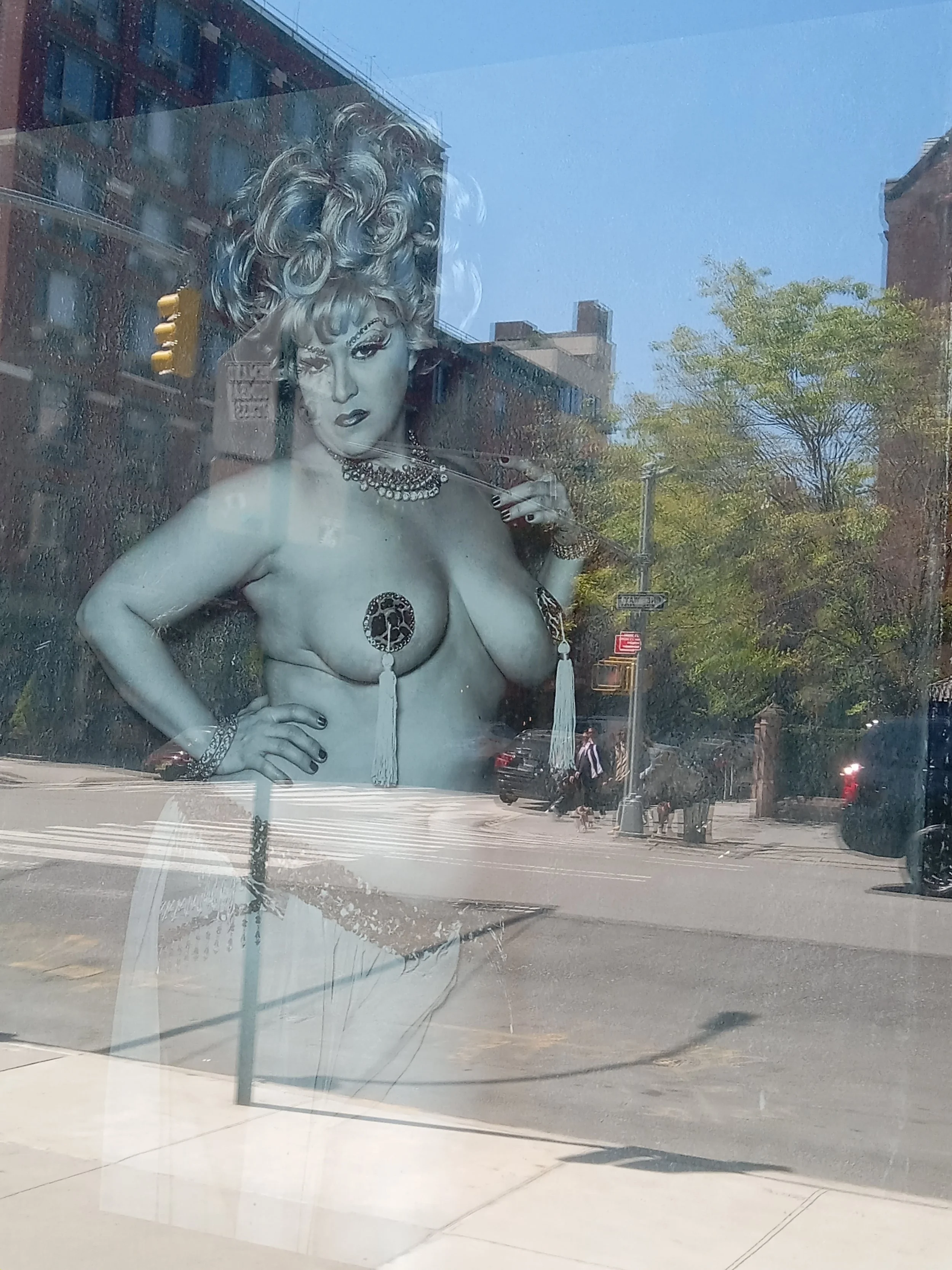 Reflection of a woman with blonde curly hair, jewelry, and a serious expression in a store window, with city street and buildings visible outside.