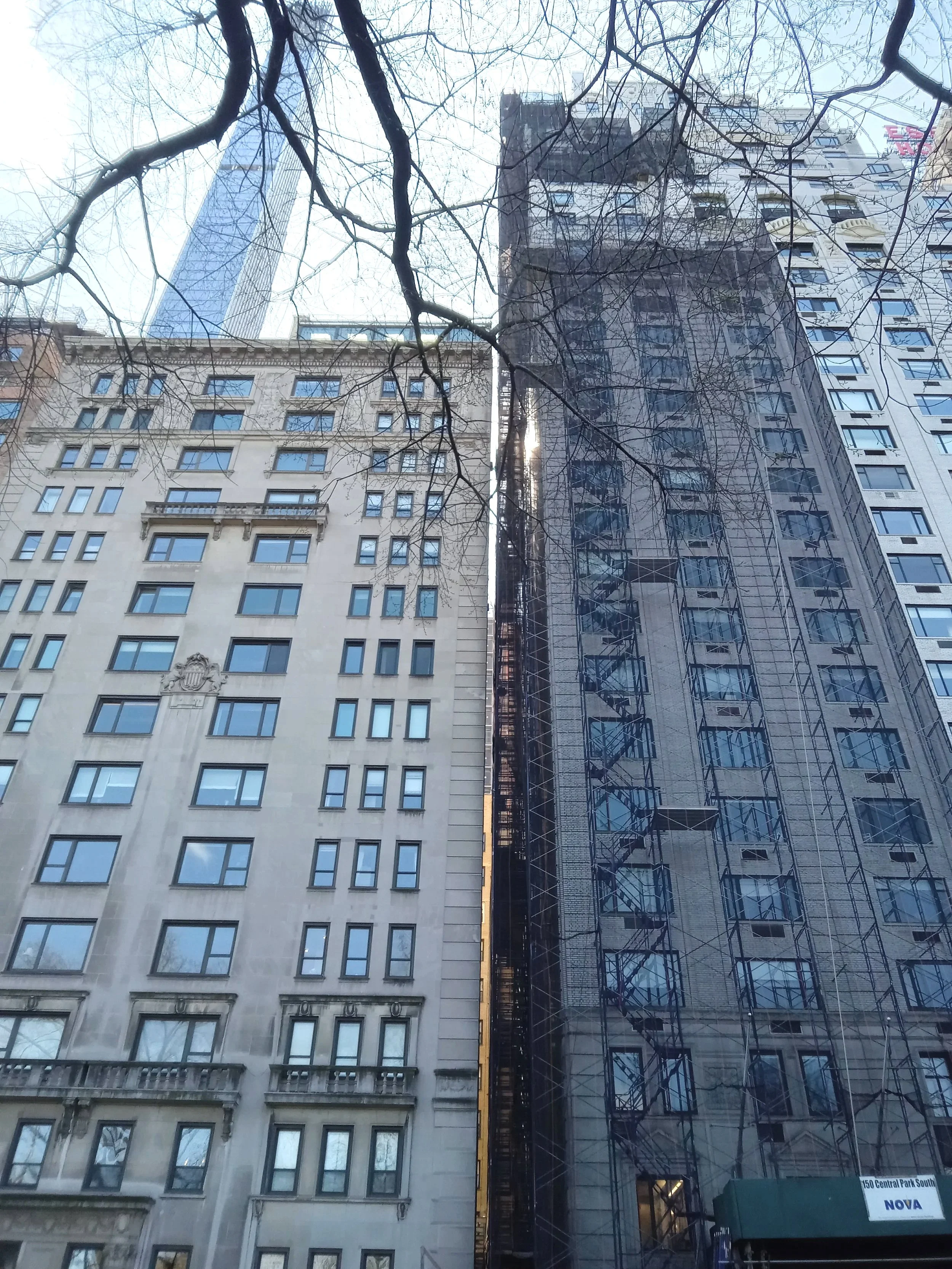 Tall buildings in an urban area with leafless tree branches in the foreground and a blue sky background. The image shows multiple buildings, including a glass skyscraper and older stone buildings.