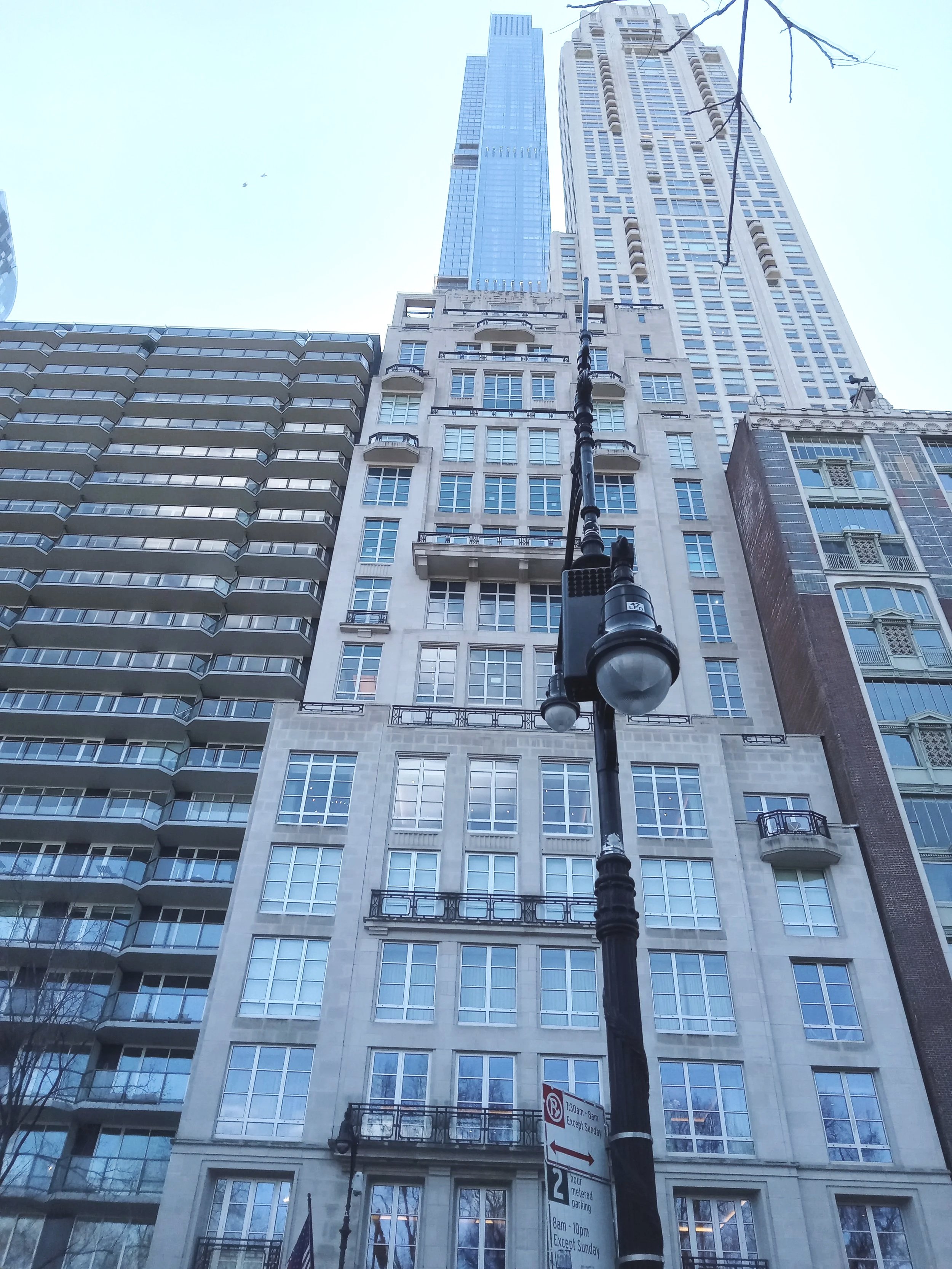 Looking up at a tall skyscraper in a city with other high-rise buildings surrounding it, under a clear sky, with a streetlamp and parking signs in the foreground.