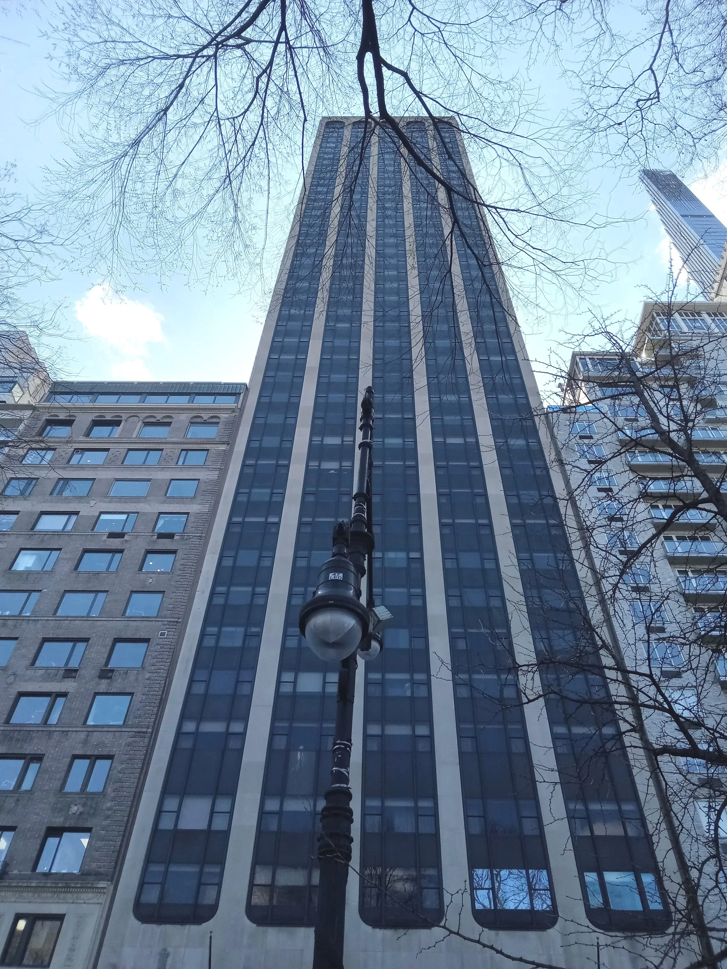 A tall skyscraper viewed from the street, with leafless tree branches surrounding it and a vintage-style streetlamp in the foreground.