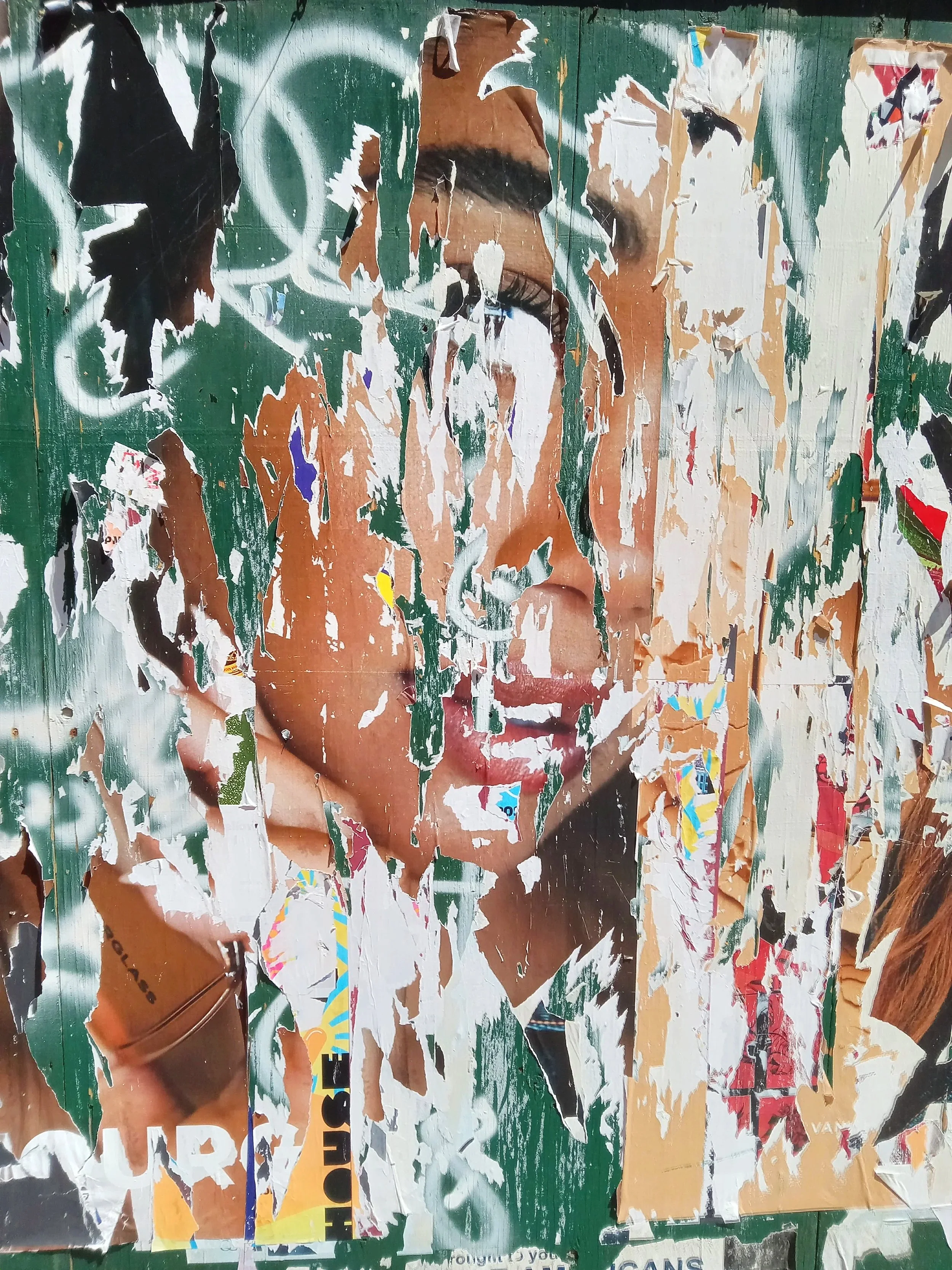 A heavily torn poster featuring a woman with dark skin and long dark hair. The poster has multiple layers of ripped paper and graffiti tags on a green background, making it difficult to see details clearly.