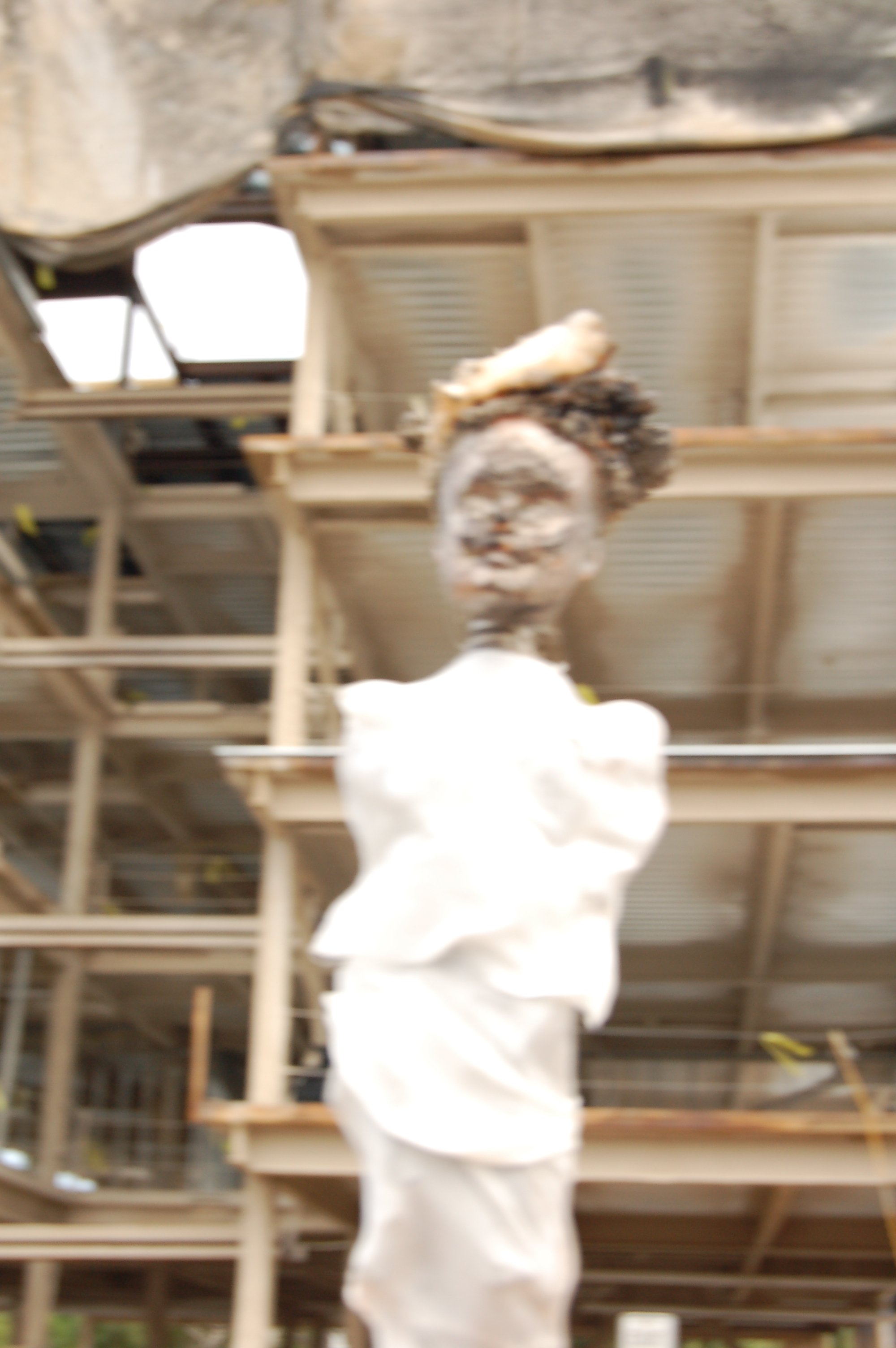 Blurred image of a person with curly hair in a white shirt in a construction or warehouse setting with metal and wooden structures.