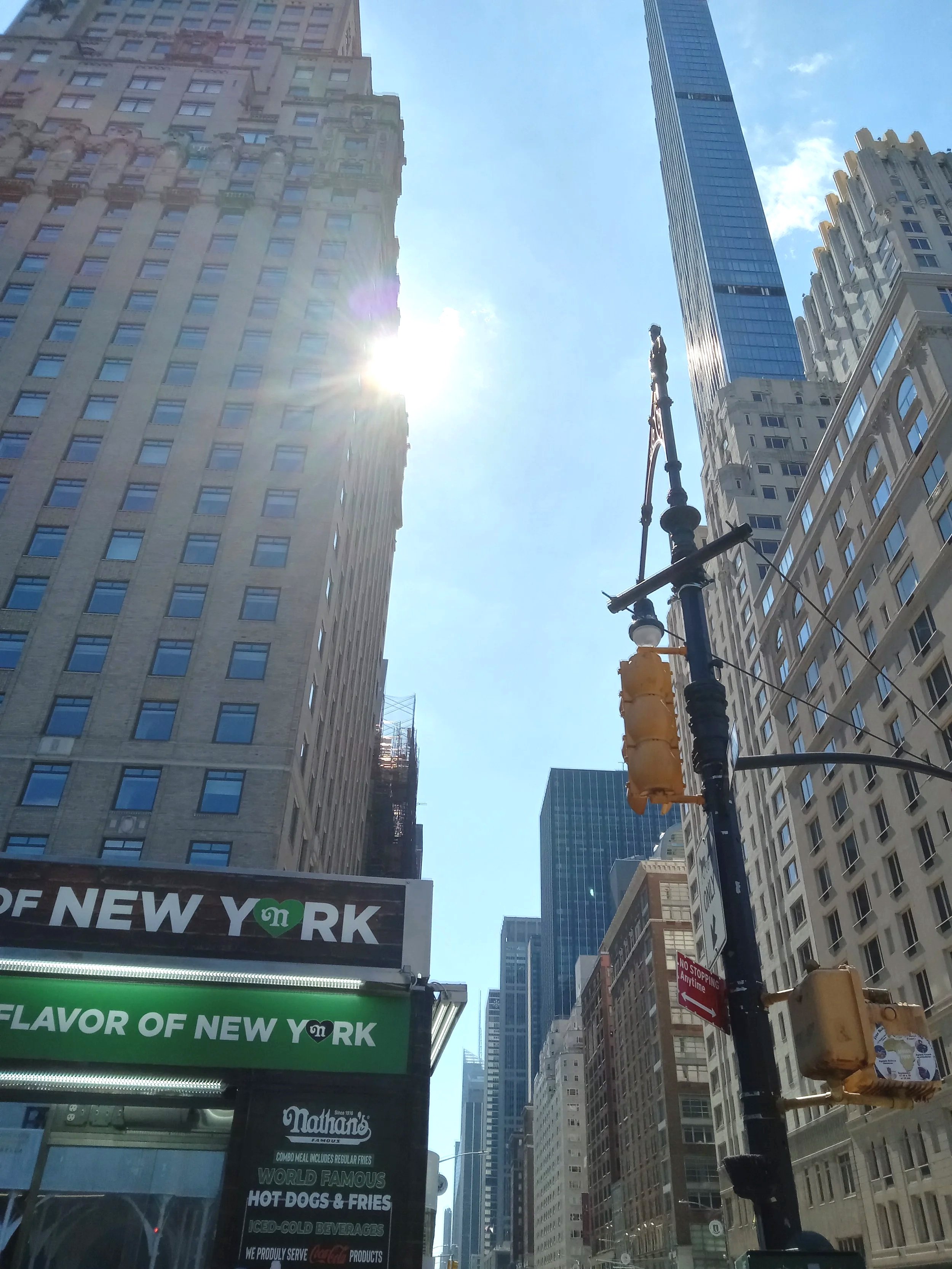 A city street scene with tall buildings, a street sign, and a traffic light pole in the foreground. The sun is shining brightly behind a building, creating lens flare. A billboard displays the words "Flavor of New York."