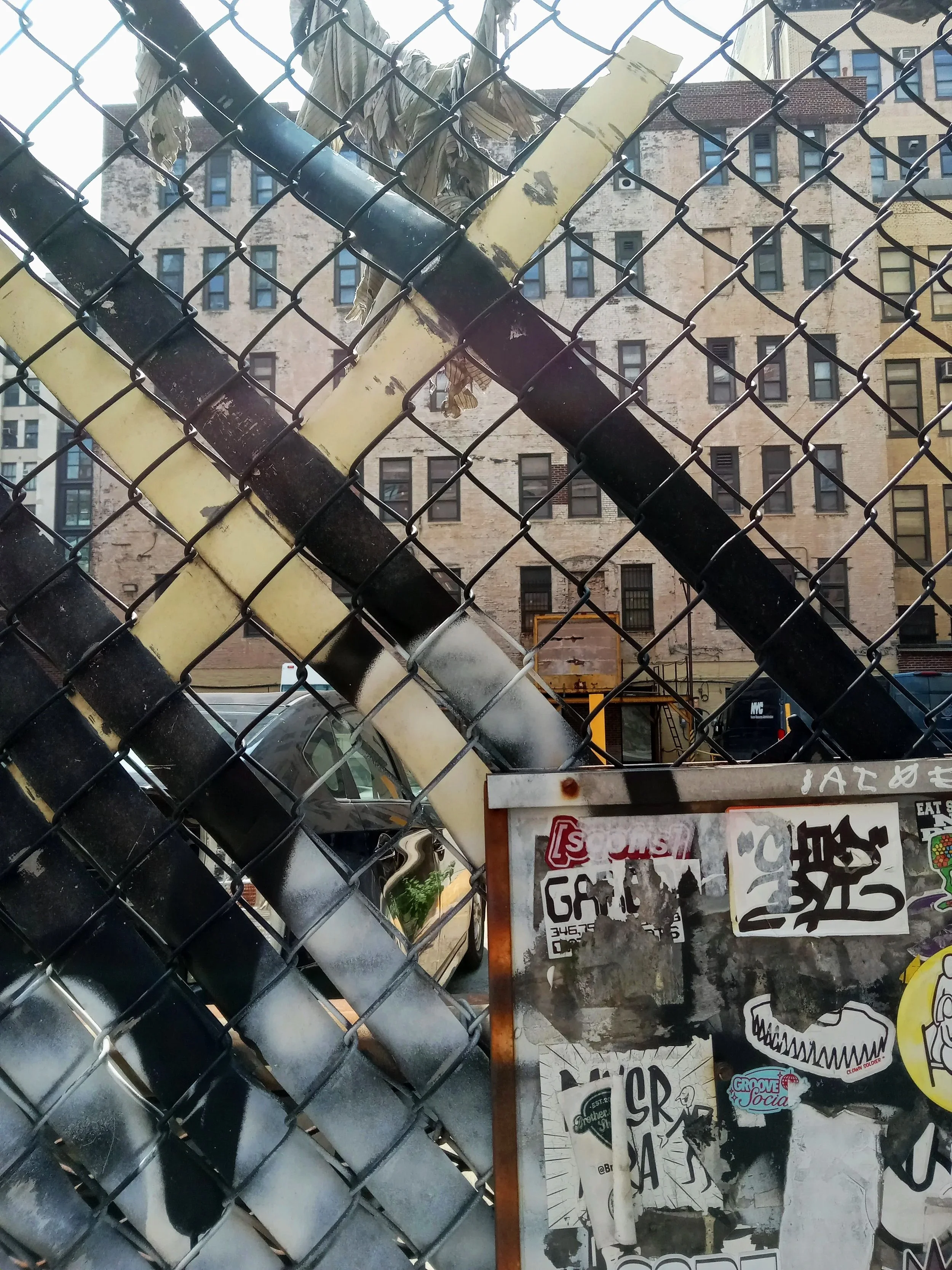 A chain-link fence with black and yellow painted pipes crossing behind it, with a city building in the background and a bulletin board with various stickers and posters attached.