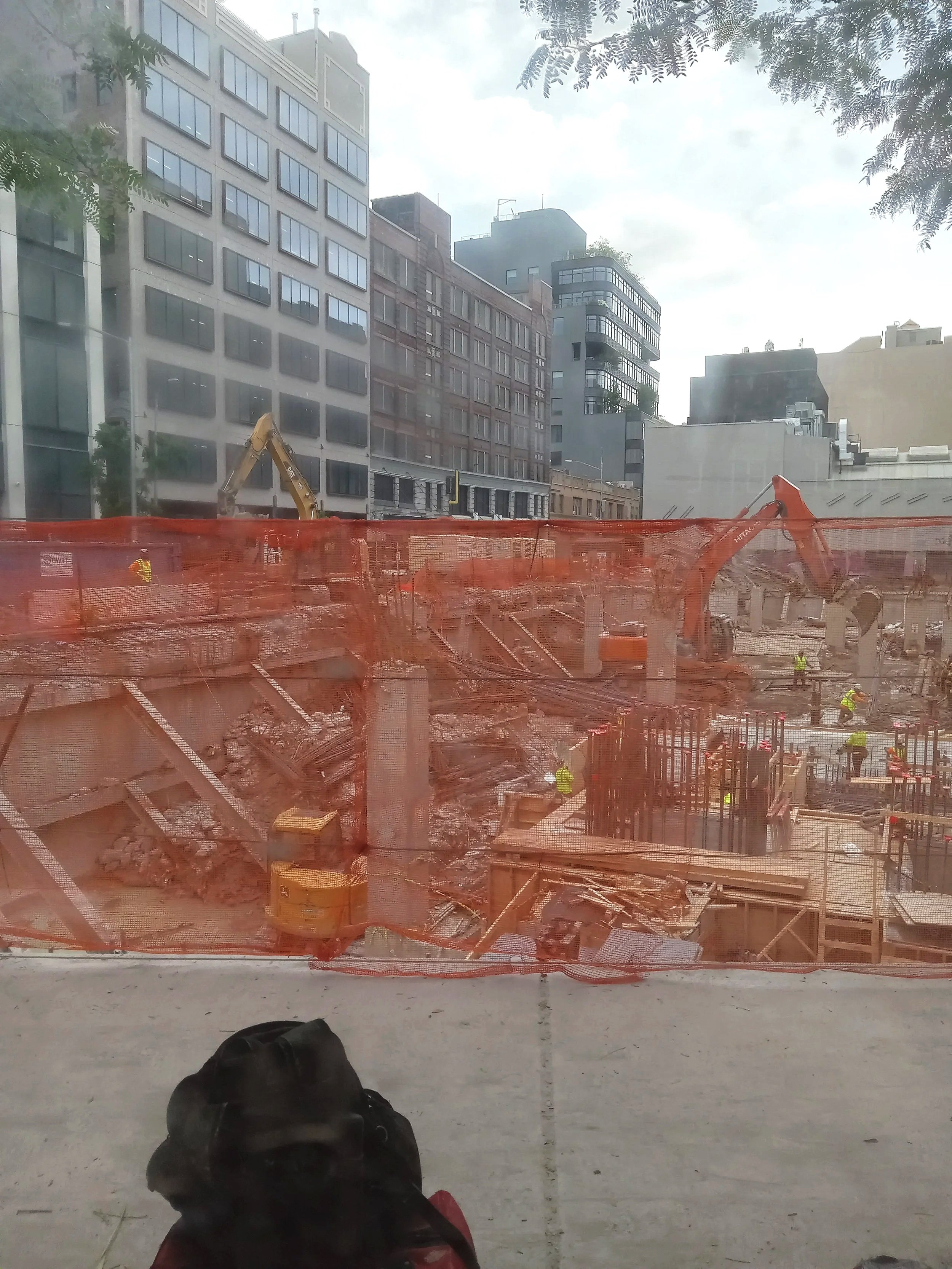 View of construction site with orange safety fence, construction workers, and machinery in front of tall buildings, seen through a window with some tree branches visible at the top.