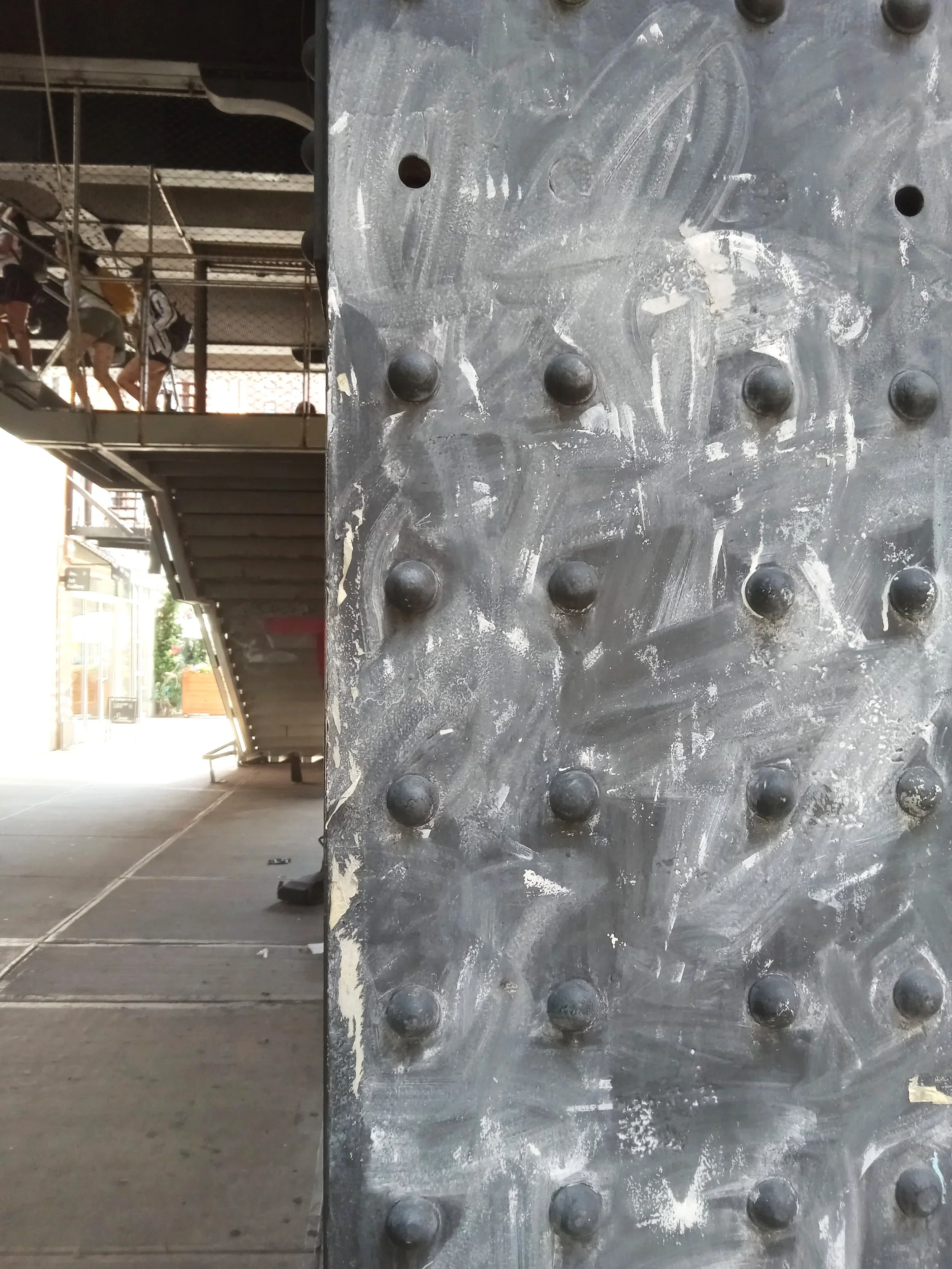 Close-up of a metallic surface with protruding round rivets, with a reflection of people walking, stairs, and a building in the background.
